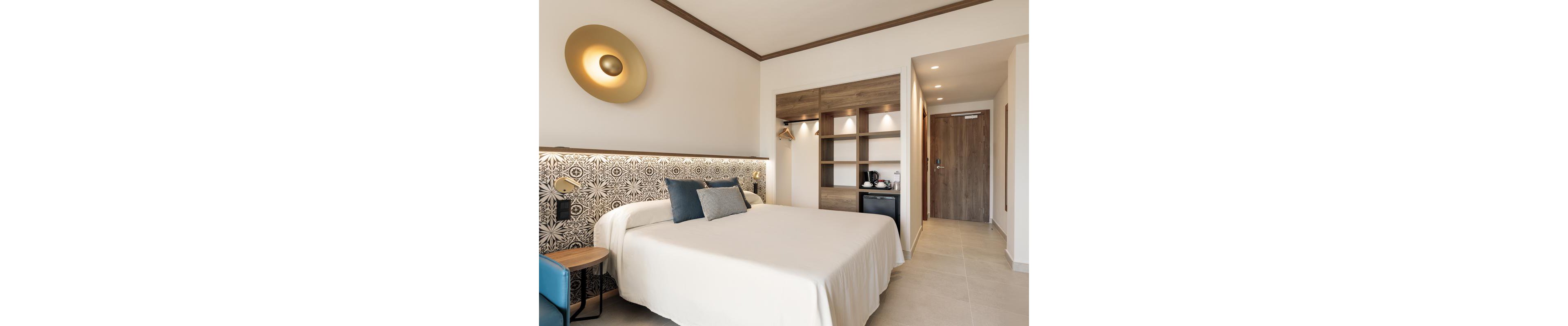 A contemporary hotel room features a neatly made bed with white linens and blue accent pillows. The setting includes a patterned headboard, wall-mounted lighting, and an open wardrobe with shelves, a mini fridge, and a coffee maker. The space is illuminated by natural and artificial light, with beige and wood tones creating a calm, inviting atmosphere. No visible text or numeric values are present in the image.