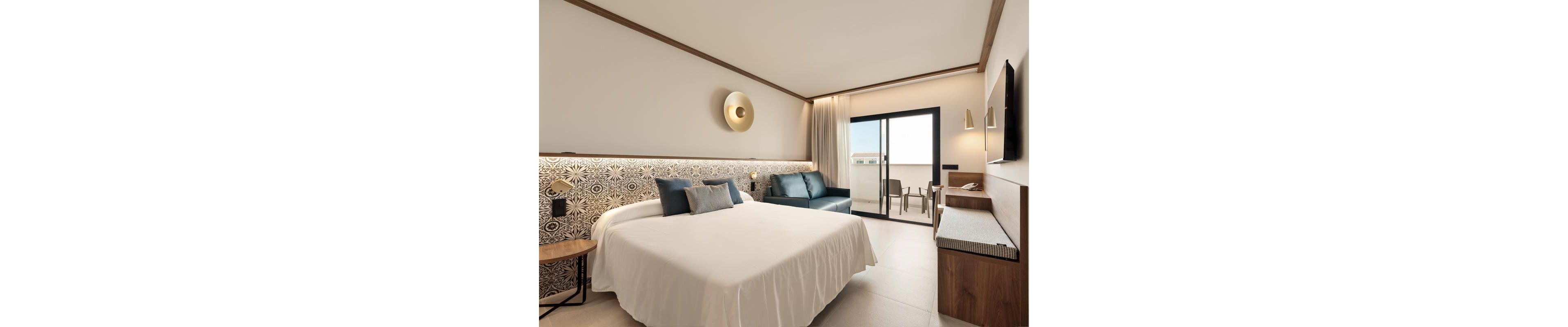 A contemporary hotel room features a large bed with patterned headboard, accent pillows, and a sleek wall lamp. The space includes a blue sofa, a wooden side table, and a flat-screen TV mounted opposite the bed. Natural light fills the room through a glass door leading to a balcony with outdoor seating, creating a bright and inviting atmosphere.