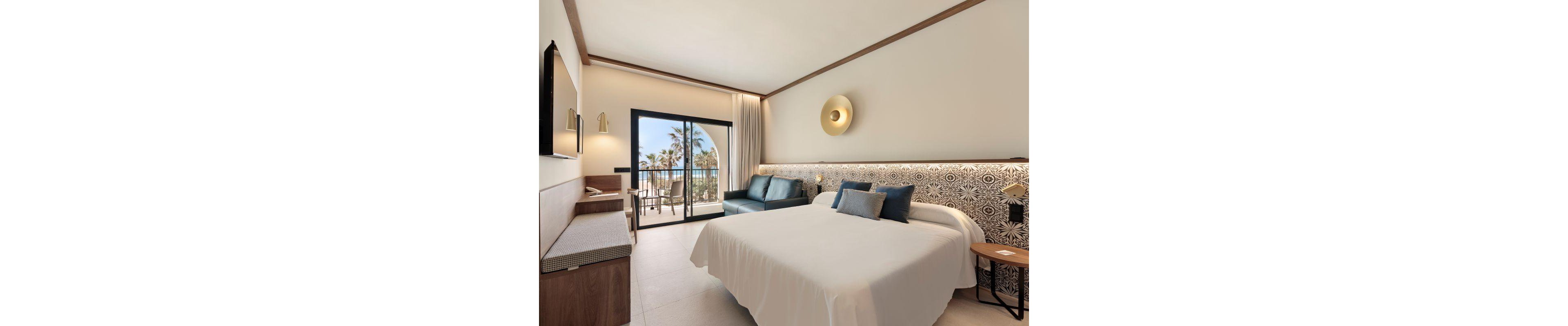 A spacious hotel room features a large bed with decorative pillows, a blue sofa, and a work desk. The room opens to a balcony with a view of palm trees and the ocean, creating a relaxing atmosphere. The decor is modern with neutral tones, geometric patterns on the headboard, and a circular wall light above the bed. Natural light fills the space through the sliding glass door.