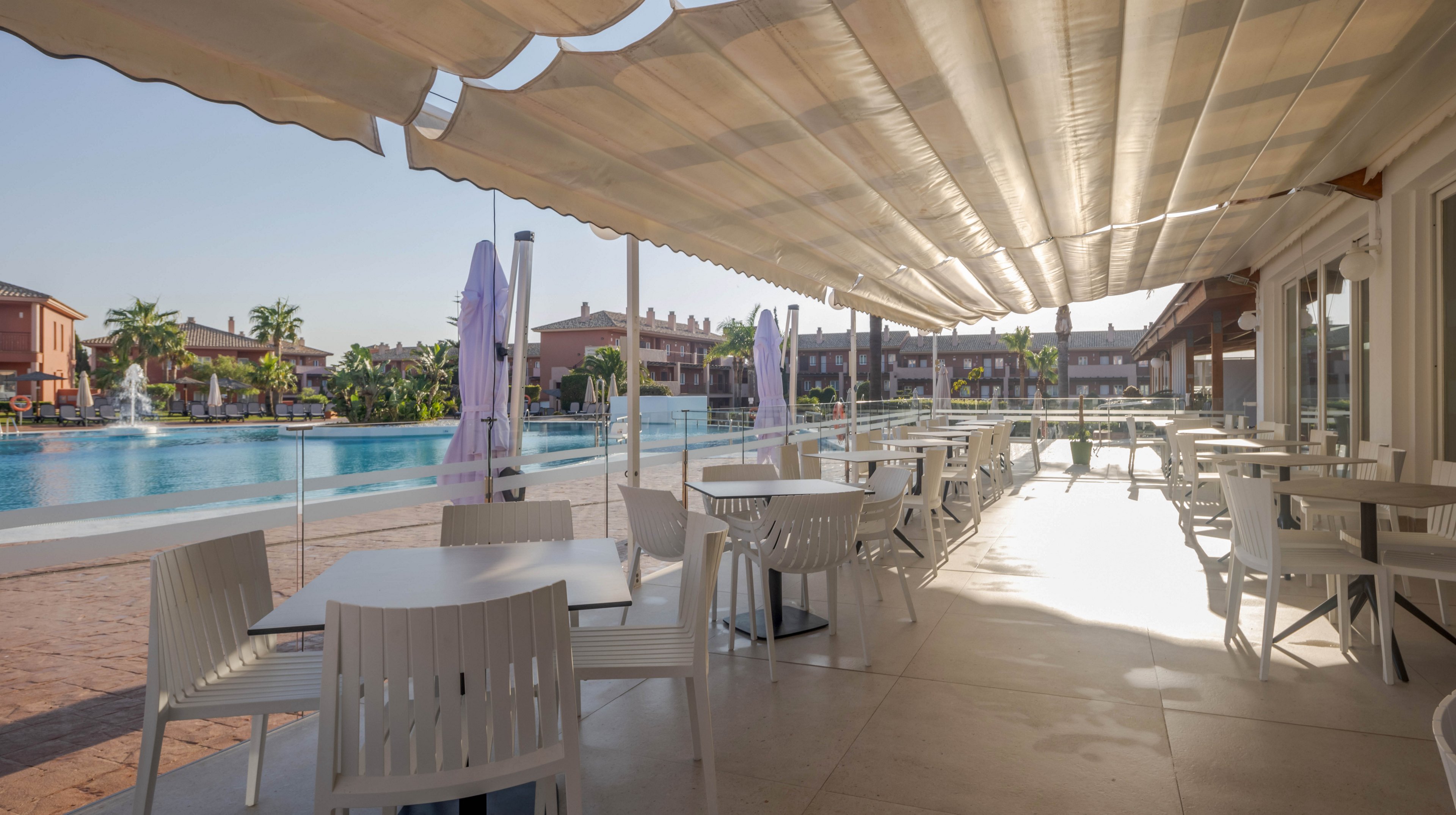 A spacious outdoor dining area features white tables and chairs under a large canopy, overlooking a clear blue swimming pool. The setting is sunlit, with shadows cast by the awning, and resort buildings are visible in the background. The atmosphere is calm and inviting, ideal for relaxation or casual meals. No people are present, emphasizing tranquility and open space. No visible text or numbers are present in the image.