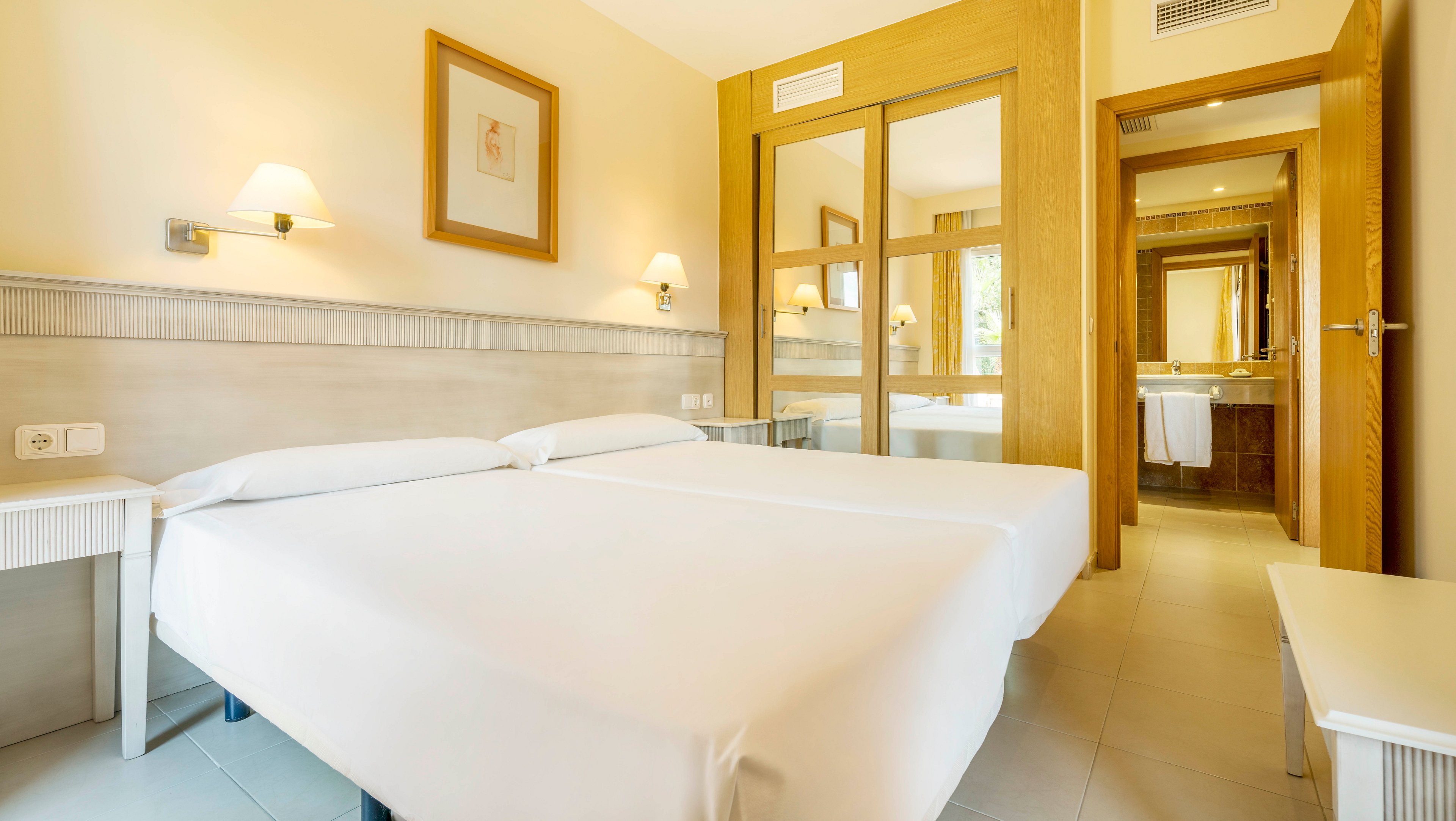 A clean, contemporary hotel room features two twin beds with crisp white linens. The space is illuminated by wall-mounted lamps and natural light reflecting off large mirrors. Light wood furniture and neutral walls create a calm, inviting atmosphere. The room includes a visible bathroom area in the background, enhancing the sense of spaciousness.