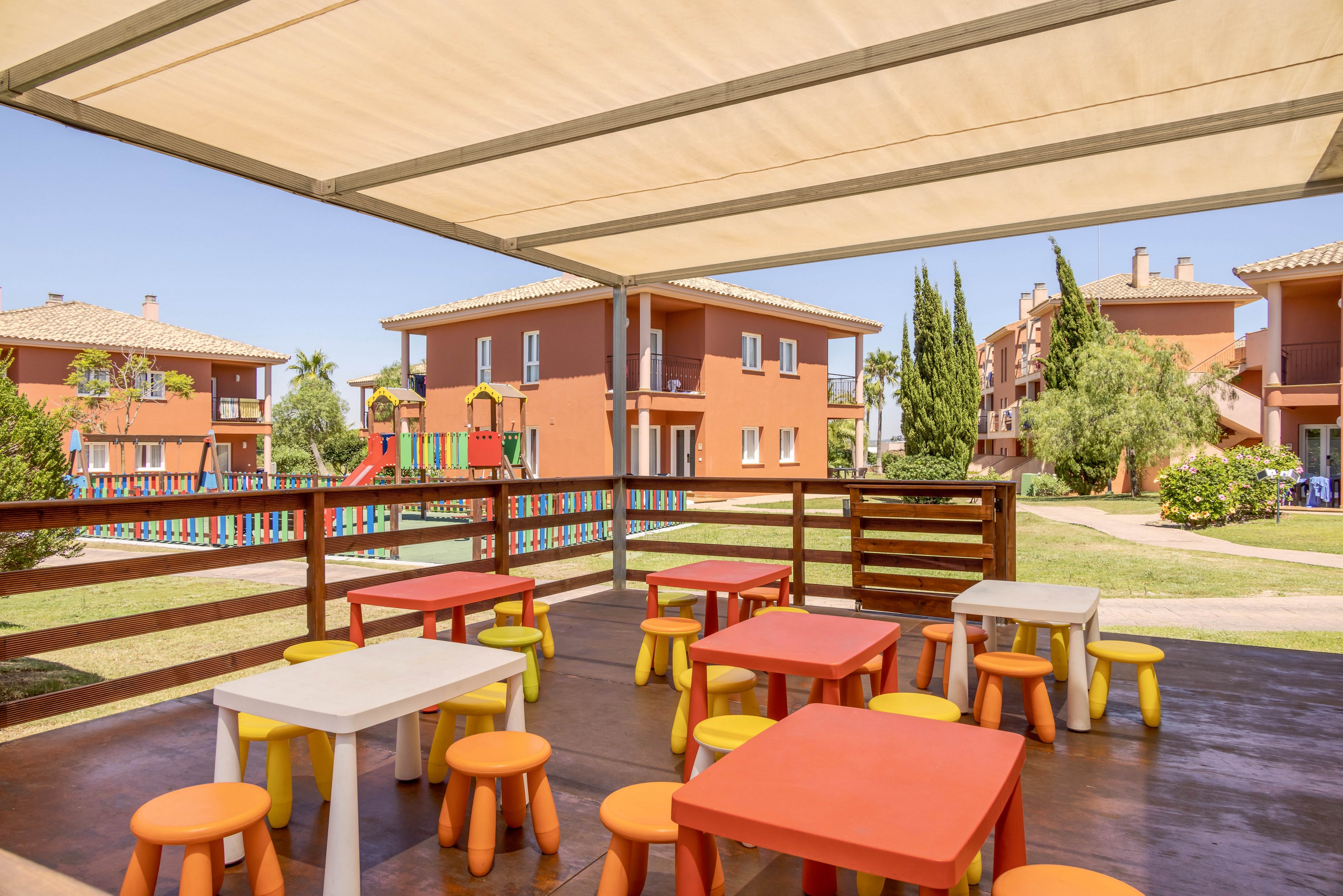 A shaded outdoor seating area features small, colorful tables and stools arranged on a wooden deck. In the background, there are red residential buildings, green lawns, and a playground with bright equipment. The scene is set on a sunny day, creating a cheerful and inviting atmosphere for children.