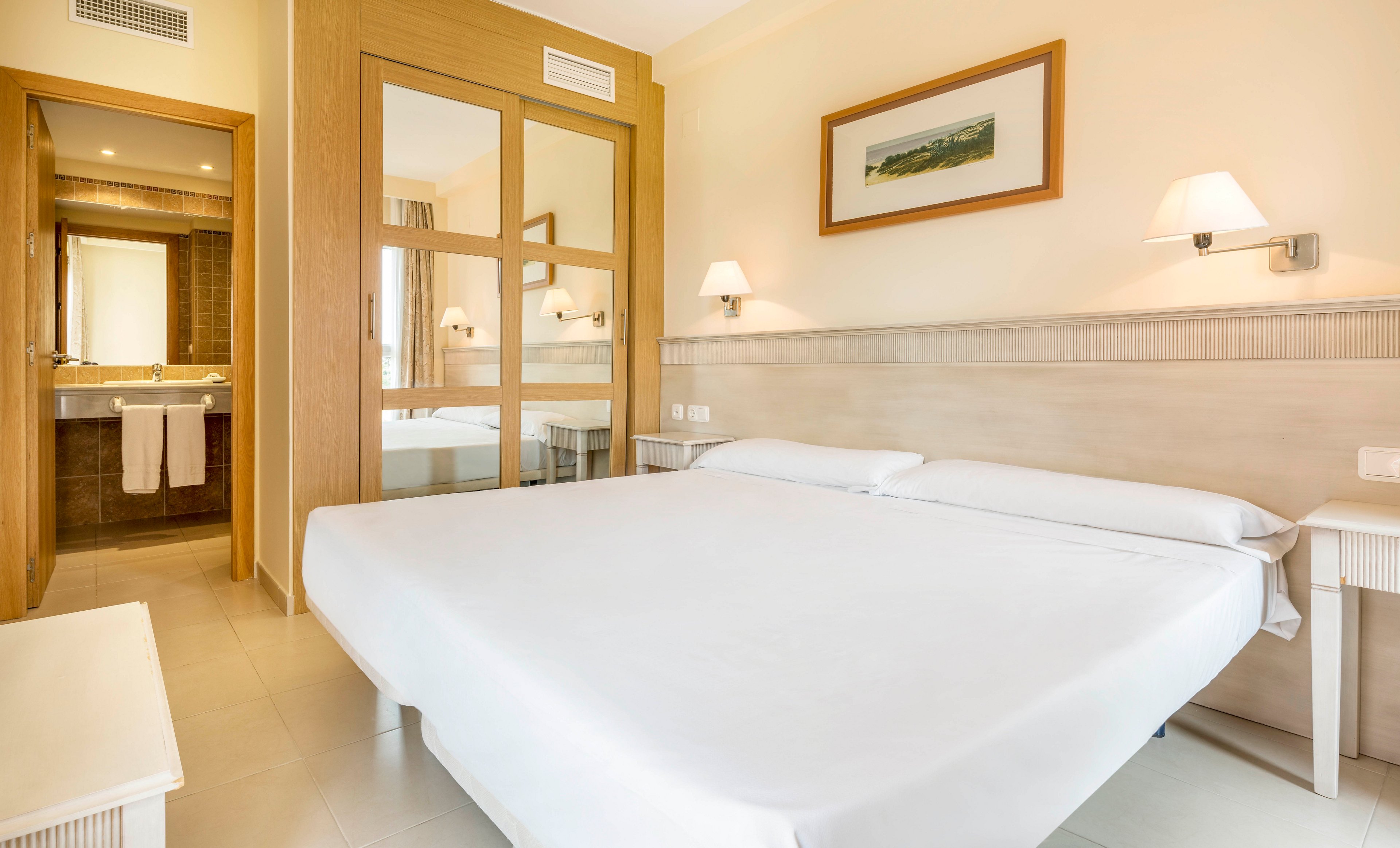 A spacious, contemporary hotel room features a large double bed with crisp white linens. The setting includes a private bathroom visible through a glass partition, with towels neatly hung. The room is decorated in neutral tones with minimalist furniture, wall-mounted lamps, and a framed landscape artwork. The overall atmosphere is clean, inviting, and well-lit.