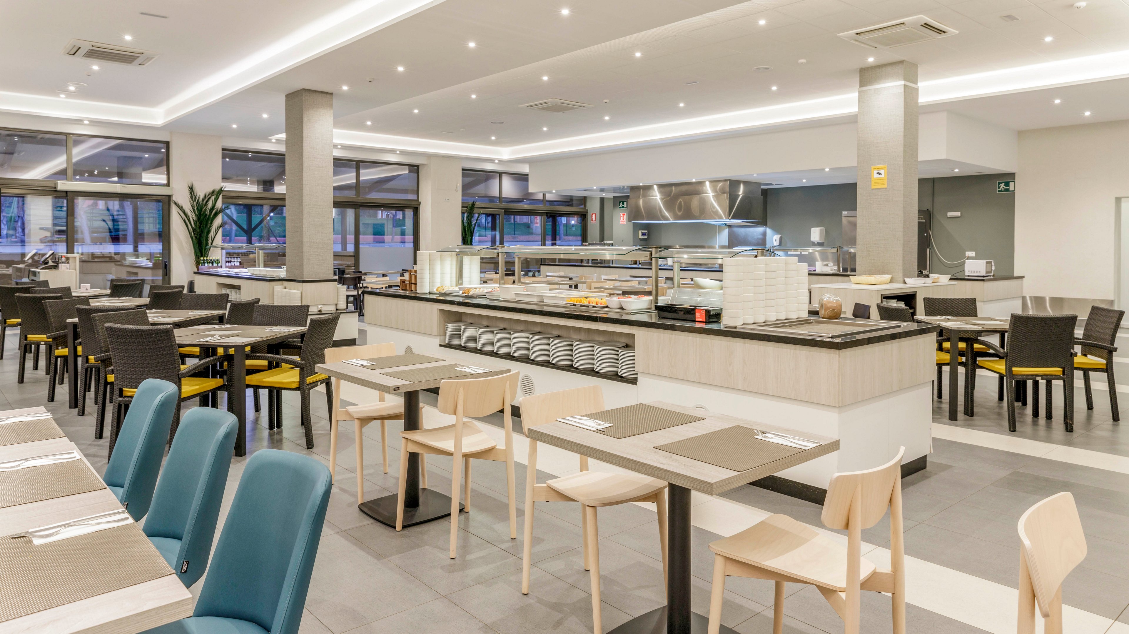 Spacious, contemporary restaurant dining area featuring a central self-service buffet counter. The setting is well-lit with recessed ceiling lights and large windows, creating an inviting atmosphere. Tables and chairs in neutral tones are neatly arranged, with a mix of seating styles. The overall palette is light and modern, emphasizing cleanliness and comfort. No visible people or explicit numeric values are present in the image.