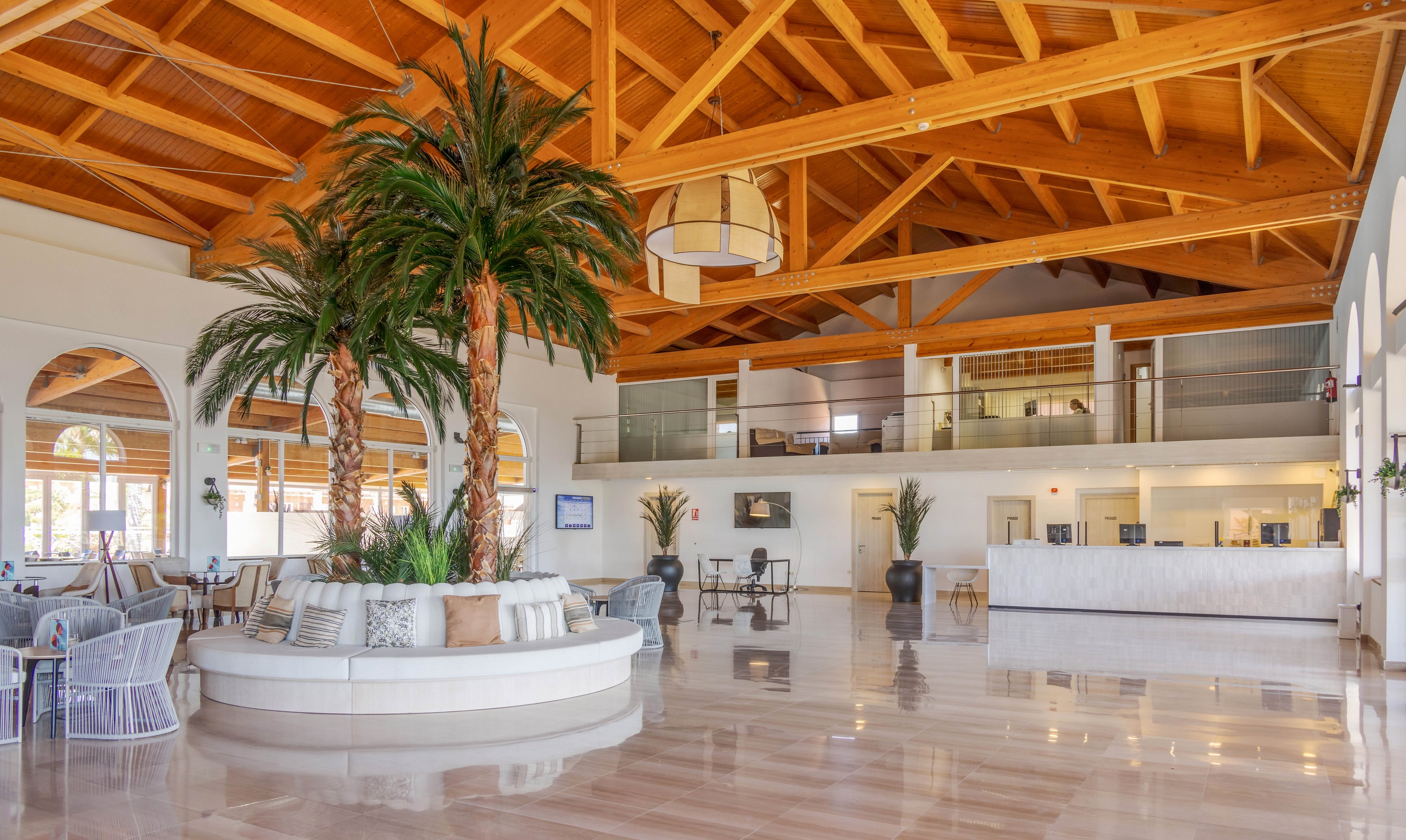 A bright and airy hotel lobby features tall indoor palm trees as a central focal point. The space is defined by high wooden ceilings, polished floors, and contemporary seating areas. Large windows and arches allow natural light to flood the room, creating a welcoming and relaxed atmosphere. The color palette is neutral with touches of greenery, and the setting appears clean and modern.