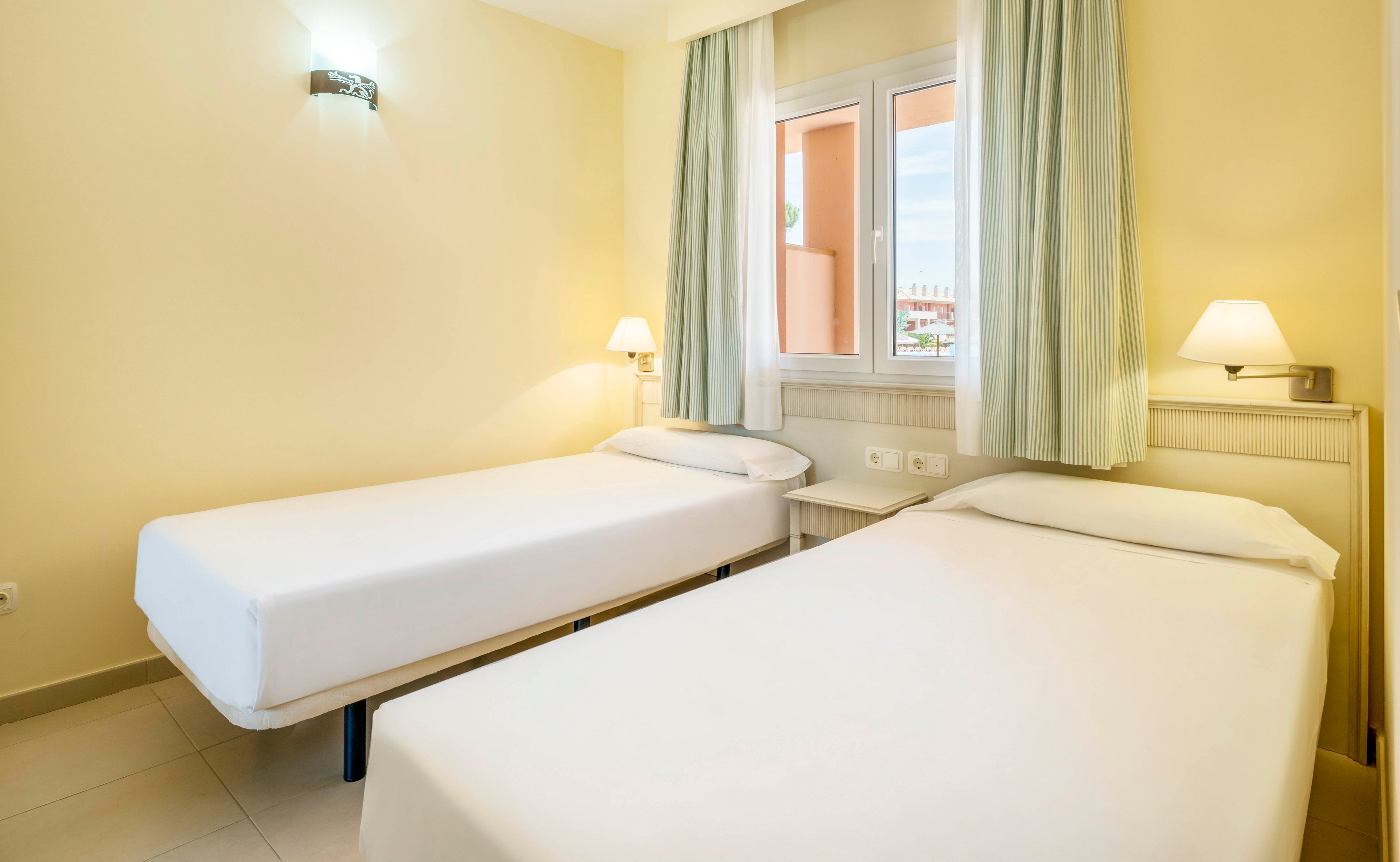 A clean, minimal hotel room features two single beds with white linens placed parallel to each other. The space is illuminated by natural light from a window with pale curtains and complemented by wall-mounted lamps. The walls are painted a soft yellow, creating a calm and inviting atmosphere. No visible text or numbers are present in the image.