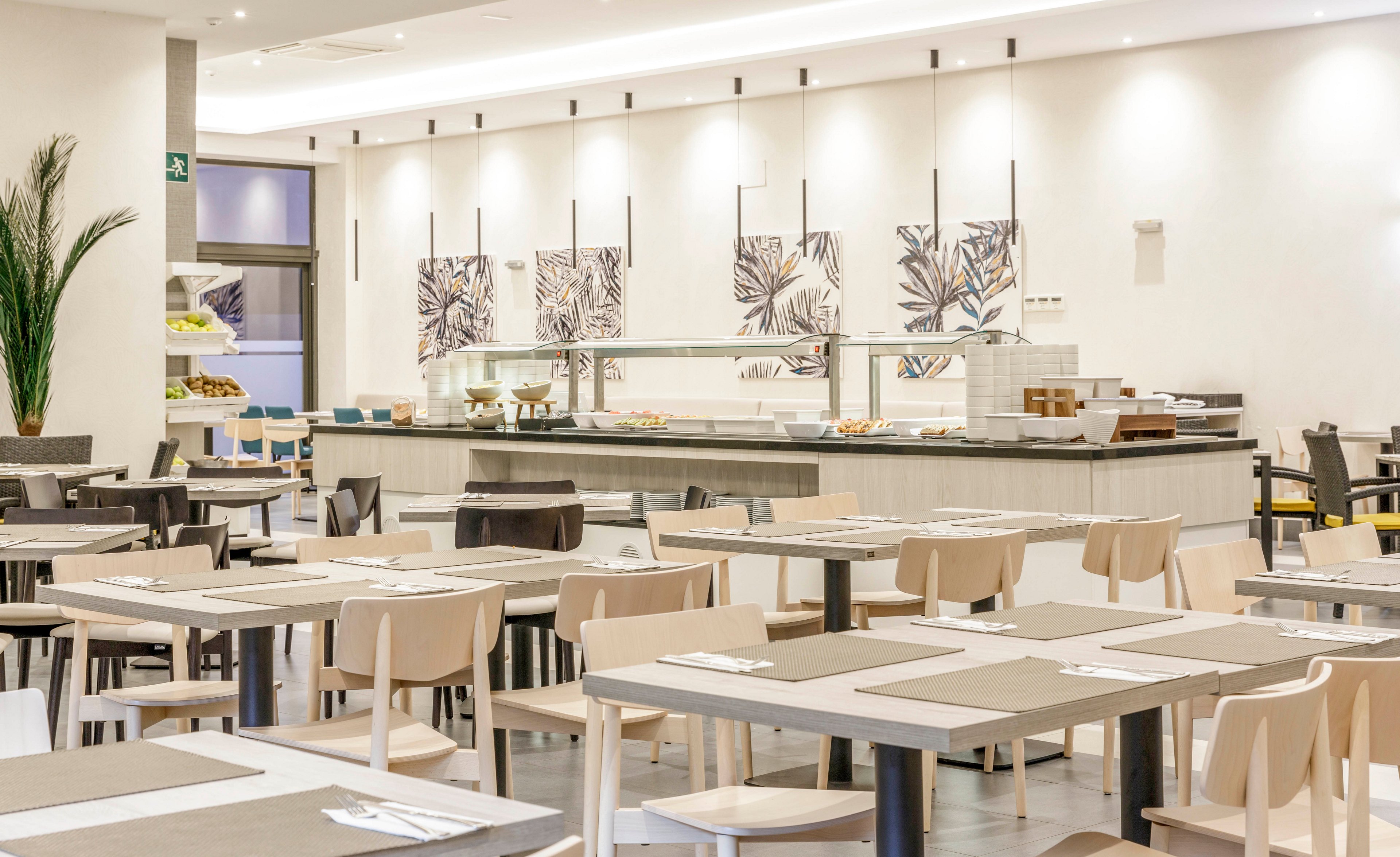 A spacious, contemporary restaurant dining area features neatly arranged tables and chairs in neutral tones. The setting includes a self-service buffet counter with food trays and decorative wall art. The atmosphere is bright and clean, with a minimalist style and soft lighting. No people are present, emphasizing the calm and orderly environment.