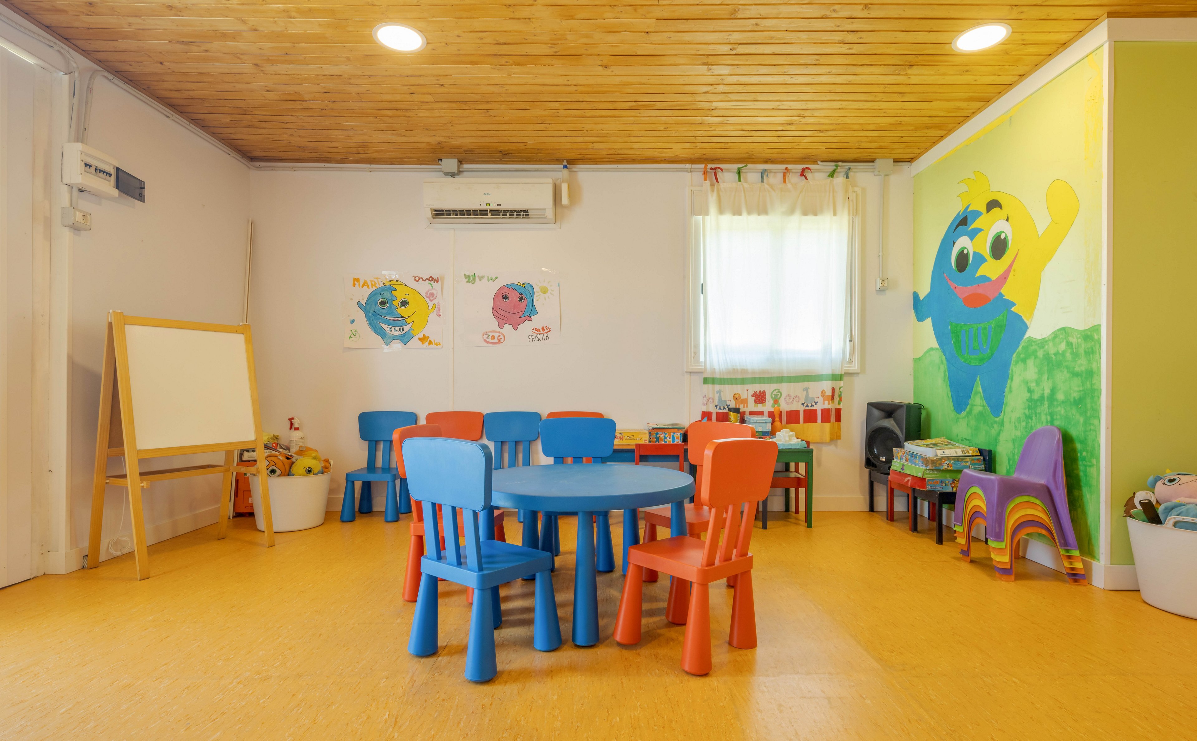 A bright and inviting preschool classroom features small blue and red chairs around a circular table. The room is decorated with cheerful cartoon wall art, including a large character mural and playful posters. Natural light enters through a window, and a wooden ceiling adds warmth to the space. An easel and educational toys are visible, creating a welcoming learning environment for young children.