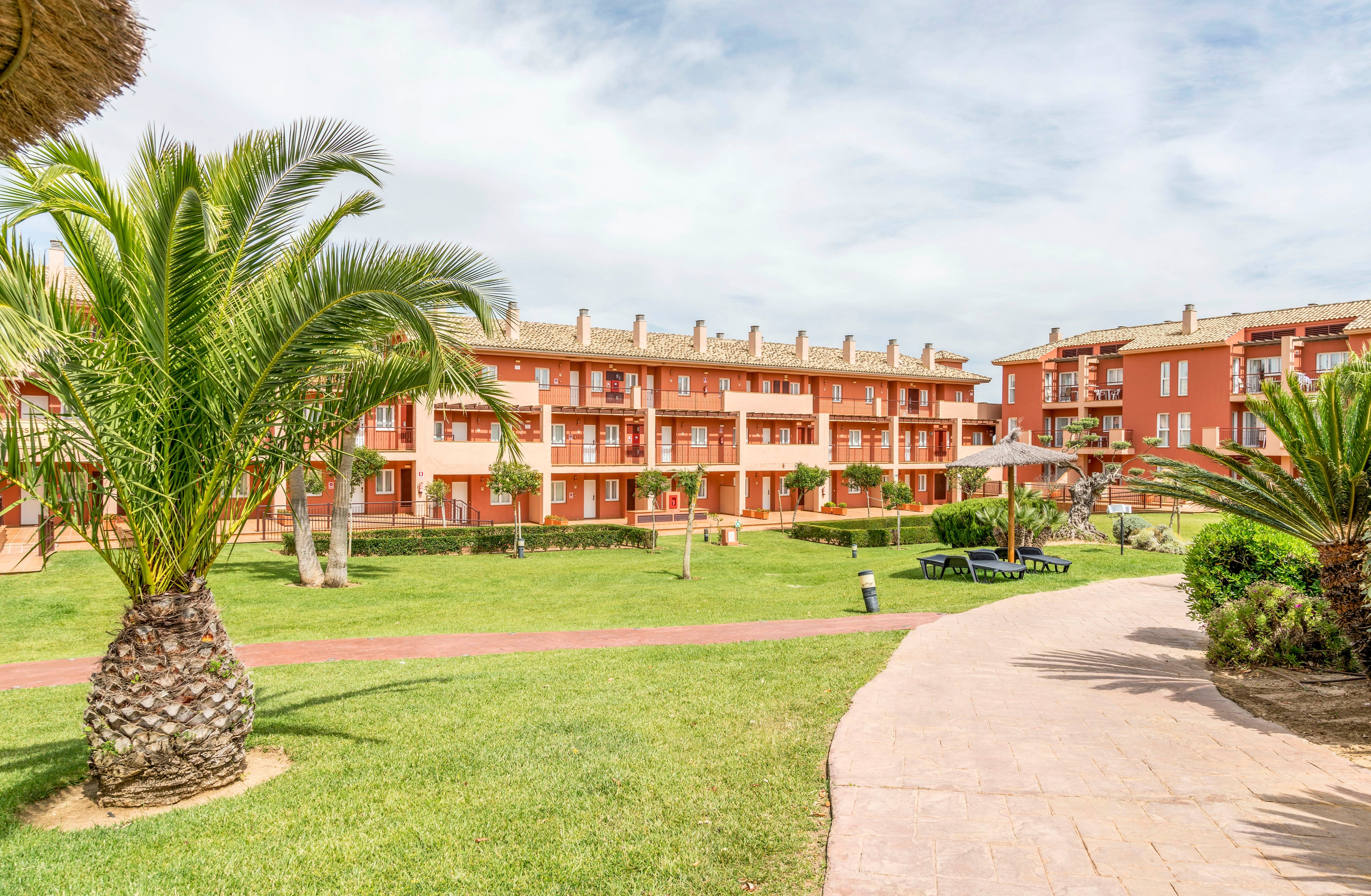 A modern apartment complex features terracotta-colored buildings surrounded by well-maintained green lawns and palm trees. The setting is outdoors on a bright, sunny day, with a paved walkway curving through the landscaped garden. Lounge chairs and umbrellas are visible, suggesting a relaxing, resort-like atmosphere. The overall mood is tranquil and inviting, ideal for leisure or vacation.