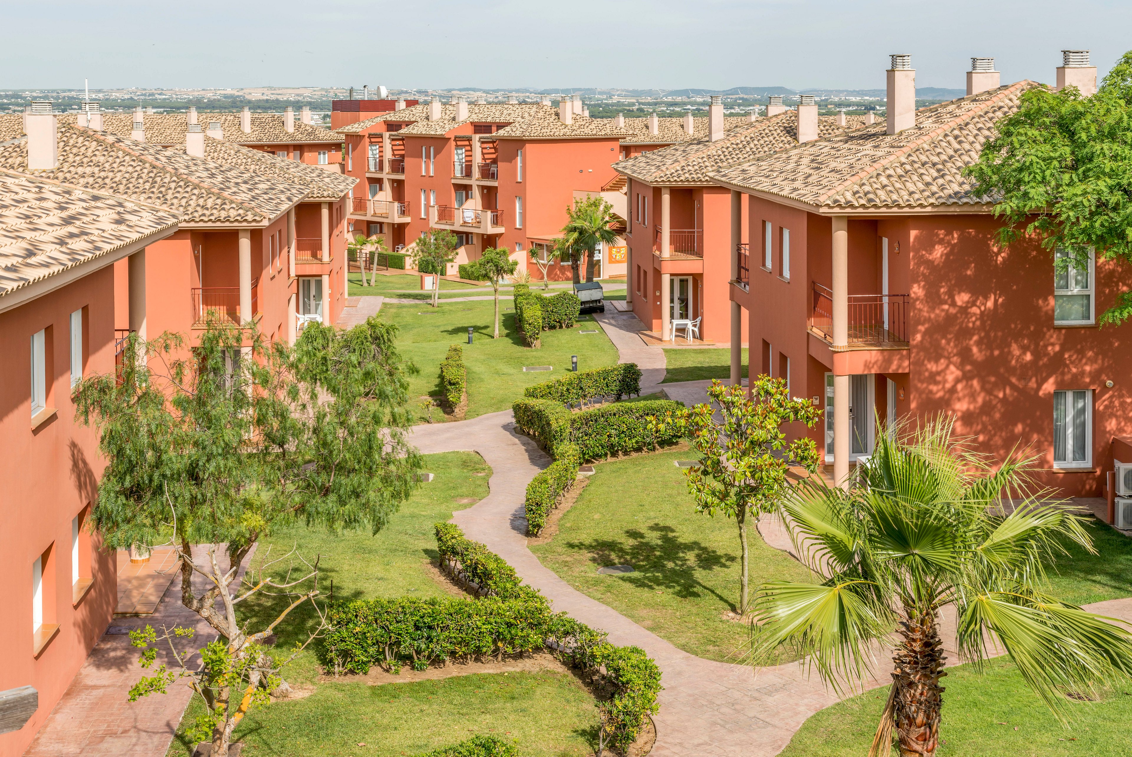 A group of terracotta-colored apartment buildings is arranged around landscaped green courtyards. The setting is bright and sunny, with well-maintained lawns, palm trees, and winding stone pathways. The architecture features tiled roofs and balconies, evoking a Mediterranean or southern European style. No people are visible, and the mood is peaceful and inviting. The image captures a suburban or resort-like residential environment.