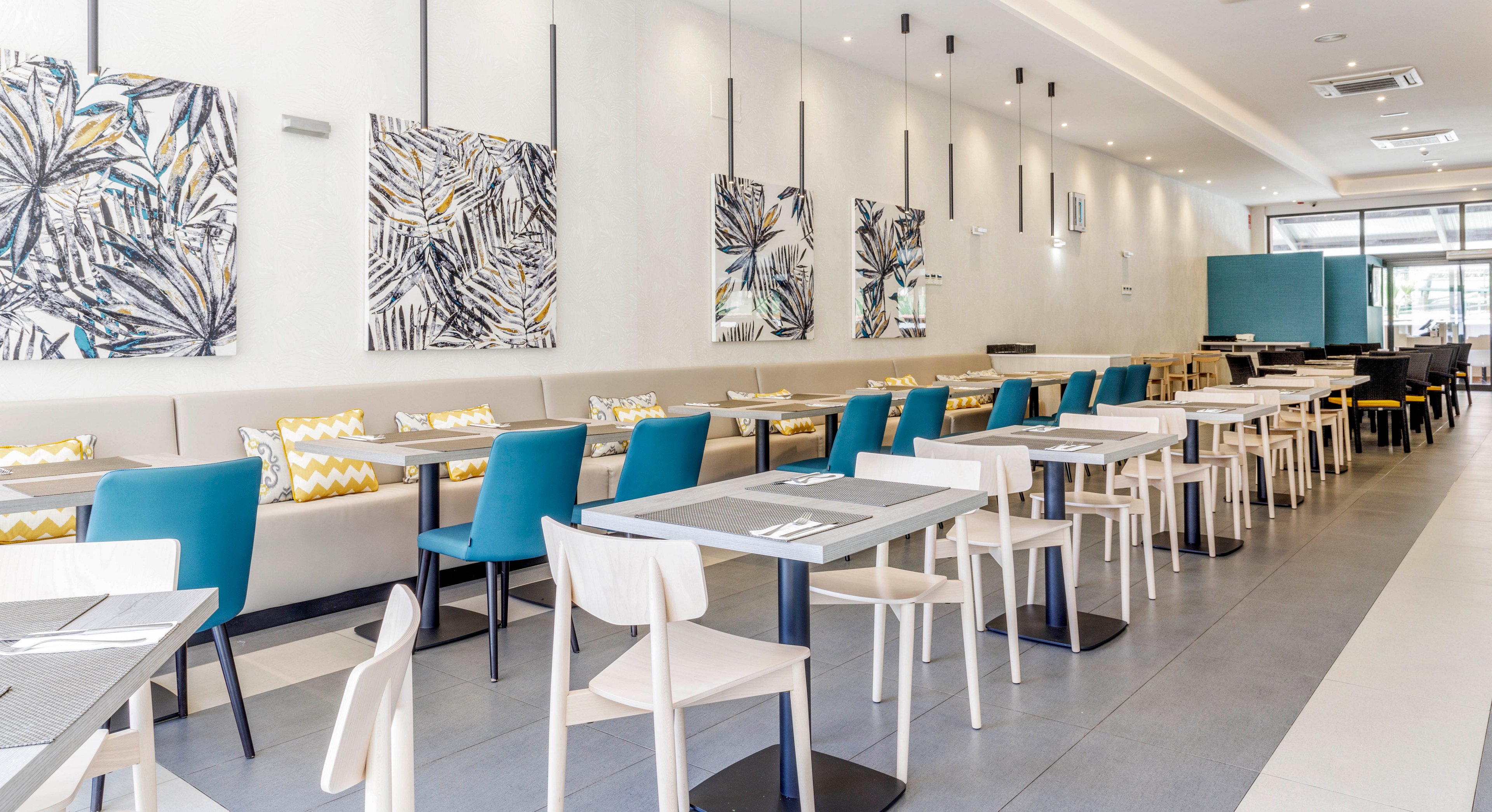 A contemporary restaurant dining area features a mix of white and blue chairs paired with sleek tables. The setting is bright and airy, with abstract botanical artwork on the walls and patterned cushions adding visual interest. The space is well-lit with pendant lights and natural light streaming in from large windows. The overall mood is inviting and stylish, with a clean, minimalist aesthetic.
