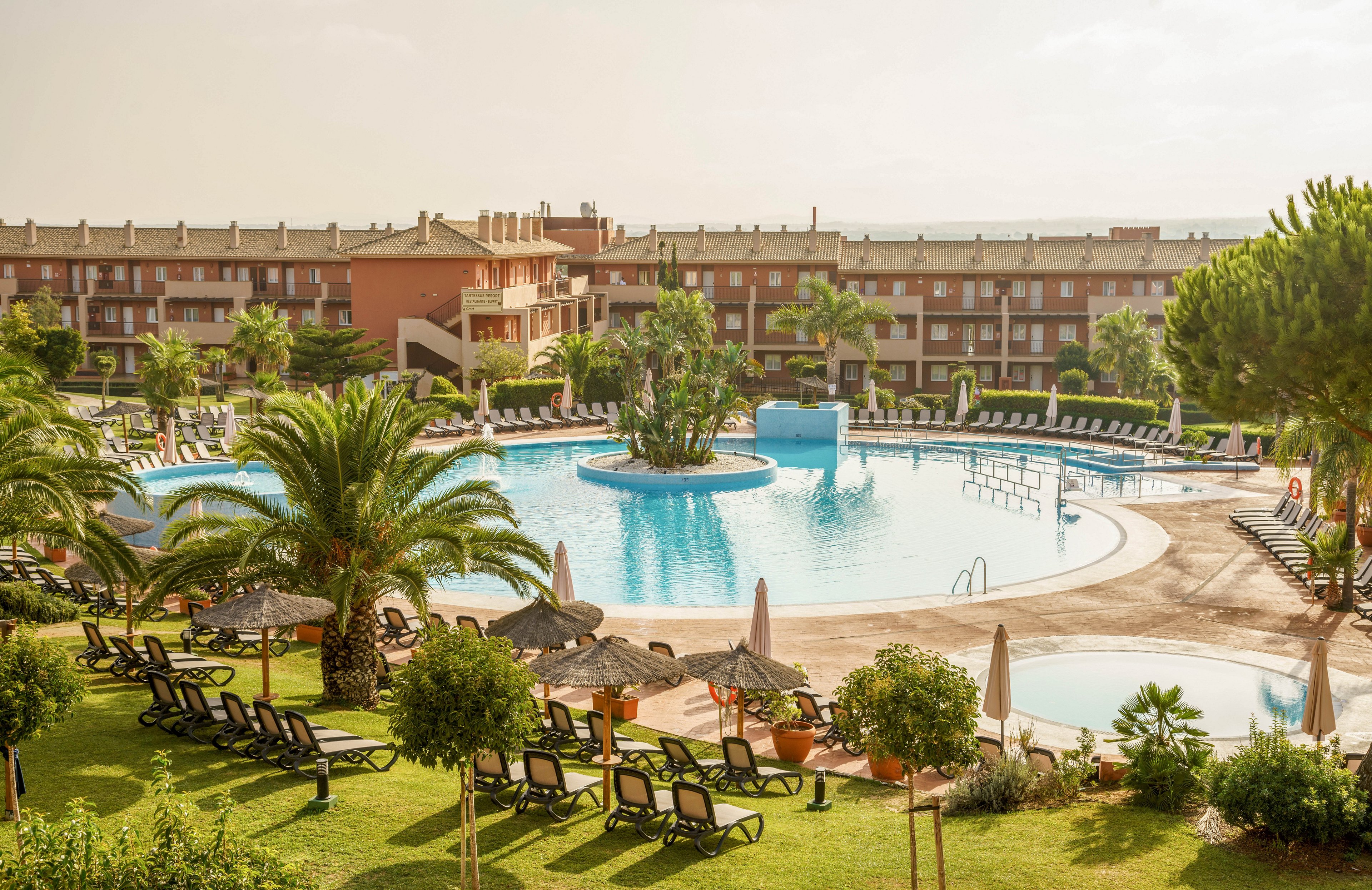 A large circular swimming pool is the central feature of this resort, surrounded by numerous black lounge chairs and lush green palm trees. The setting is outdoors with a multi-story hotel building in the background, under a bright, sunny sky. The atmosphere is relaxed and inviting, ideal for vacation or leisure. No visible text or numbers are present in the image.