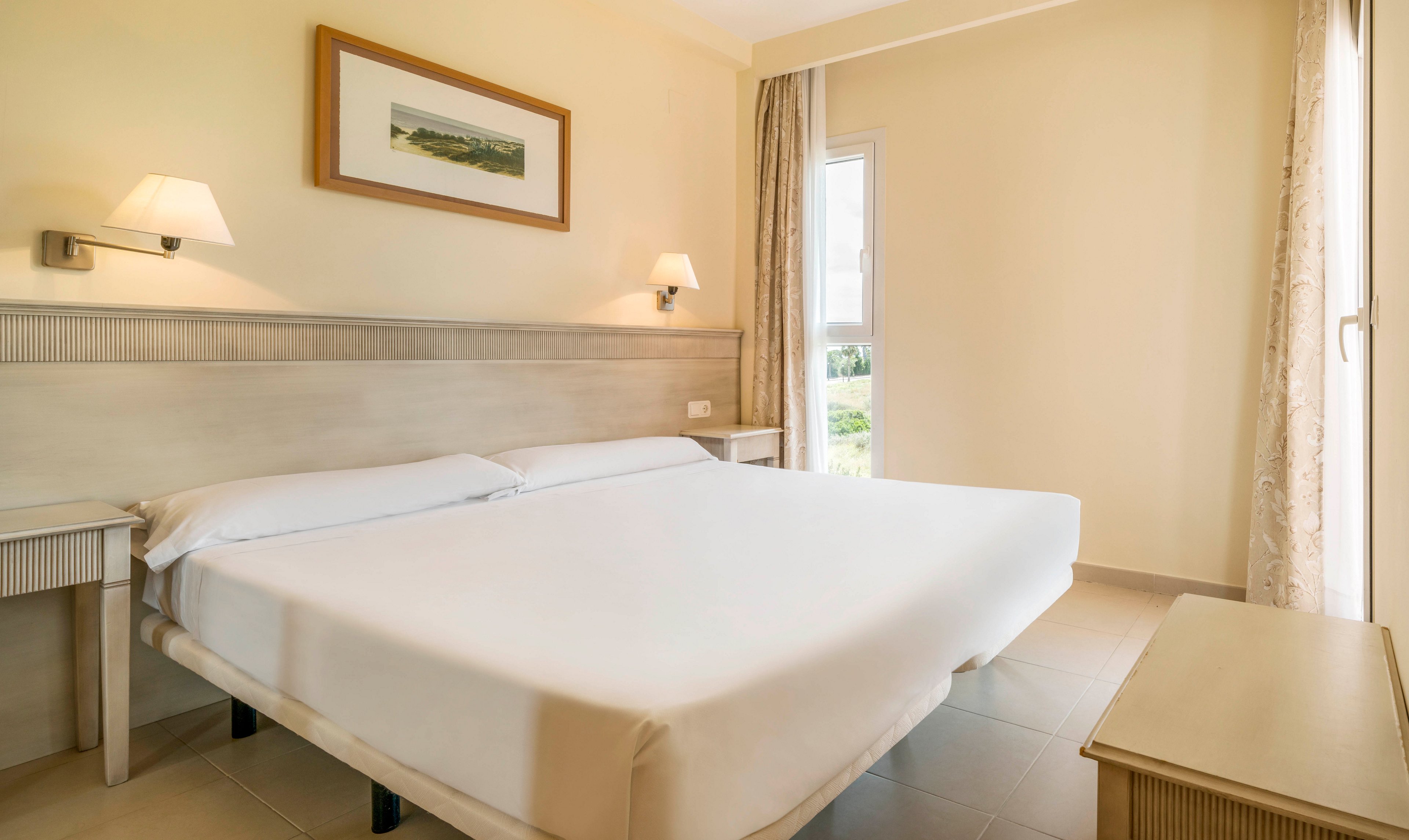 A clean and modern hotel room features a large double bed with crisp white linens. The space is illuminated by natural light from a window and complemented by two wall-mounted lamps. Neutral tones and minimalist decor create a calm, inviting atmosphere. A framed landscape artwork hangs above the bed, and simple bedside tables complete the look.