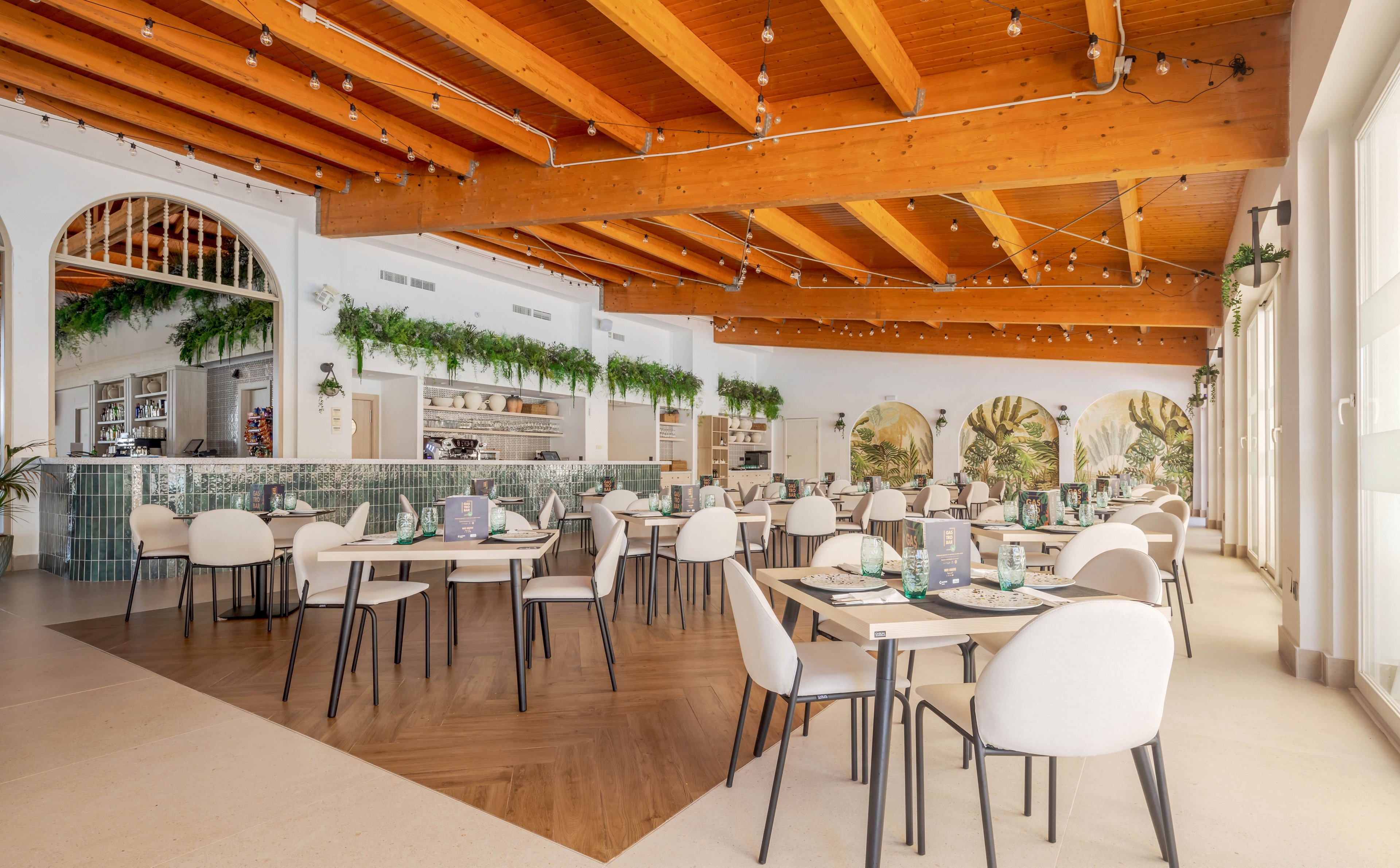 Spacious restaurant dining area featuring contemporary white chairs and tables arranged neatly. The setting includes a bar area with green tile accents, lush hanging plants, and large arched windows letting in natural light. Warm wooden ceiling beams and decorative string lights create a welcoming, airy atmosphere. No people are present, emphasizing the clean and inviting environment.