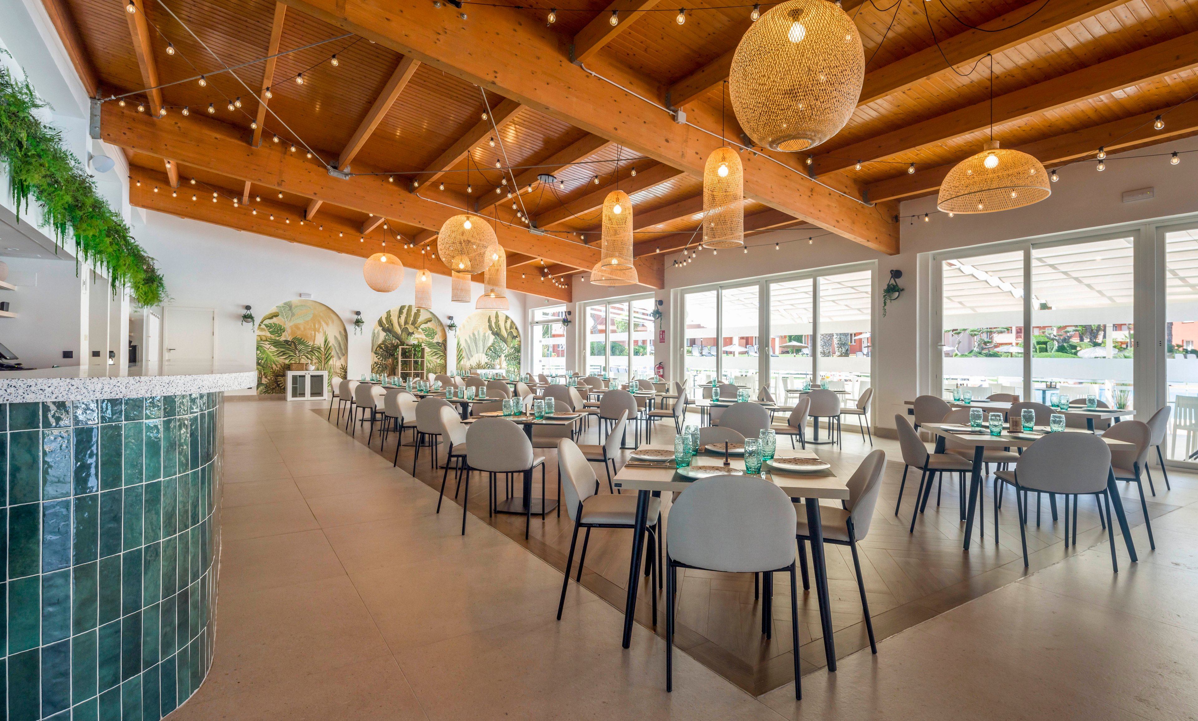 Spacious restaurant dining area featuring contemporary furniture and a high wooden beam ceiling. Warm pendant lighting and string lights create a cozy, inviting atmosphere. Large windows allow natural light to fill the space, while green tiles and wall art add visual interest. Tables are set with glassware and plates, ready for guests. No visible text or numbers are present in the image.