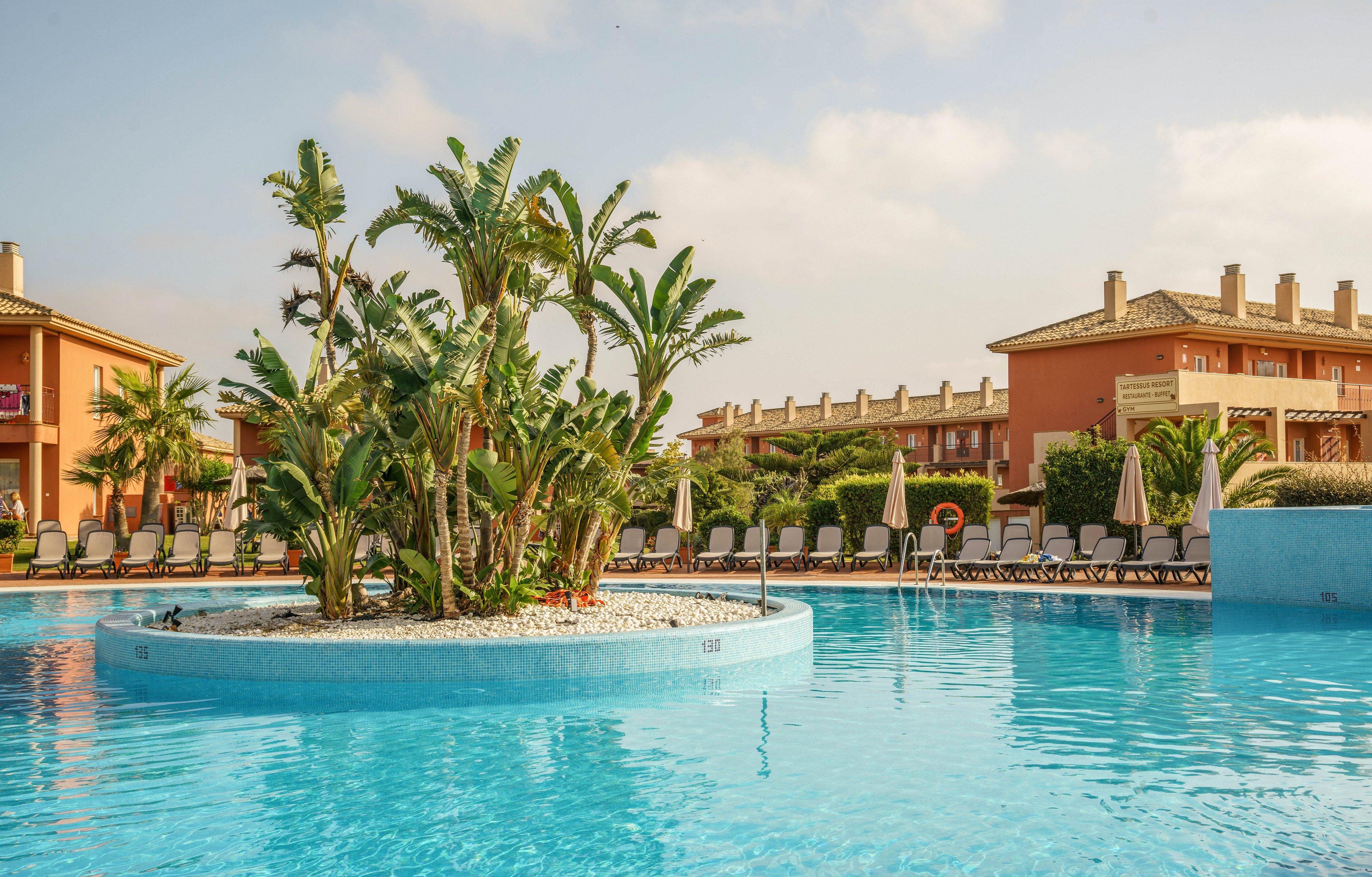 A large outdoor swimming pool features a central island with lush palm trees and white stones. Surrounding the pool are rows of sun loungers and umbrellas, set against a backdrop of Mediterranean-style resort buildings. The scene is bathed in warm daylight, creating a relaxed and inviting vacation atmosphere. No visible text or numbers are present in the image.