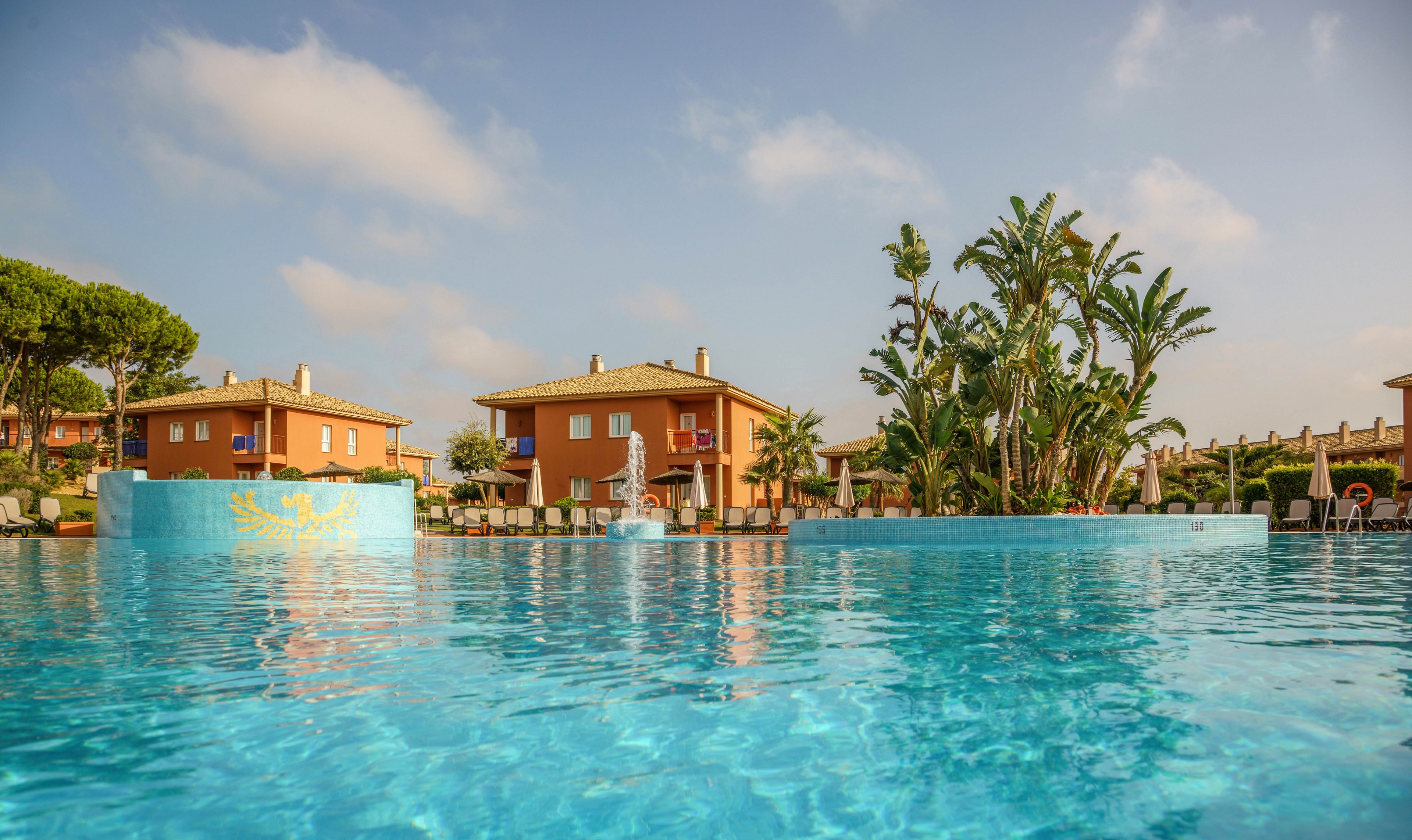 A large, inviting swimming pool is the main focus, with clear blue water reflecting the sky. In the background, there are several villa-style buildings surrounded by lush palm trees and greenery. The setting appears to be a sunny, tranquil resort, ideal for relaxation and leisure. The overall mood is calm and vacation-like, with warm tones and a bright atmosphere.