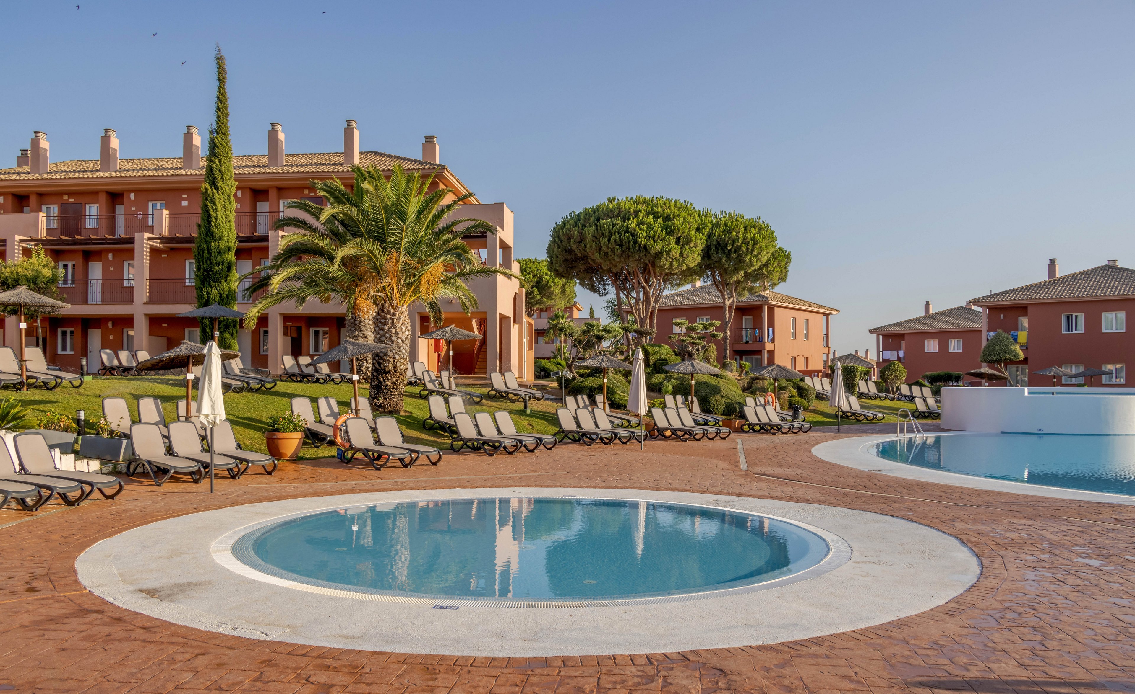 A tranquil resort pool area features a circular wading pool in the foreground and a larger swimming pool nearby. Numerous sun loungers are arranged neatly around the pools, set against manicured lawns and lush palm trees. Mediterranean-style buildings with terracotta roofs provide a scenic backdrop under a clear blue sky. The setting evokes a relaxed, vacation atmosphere with warm, natural lighting.