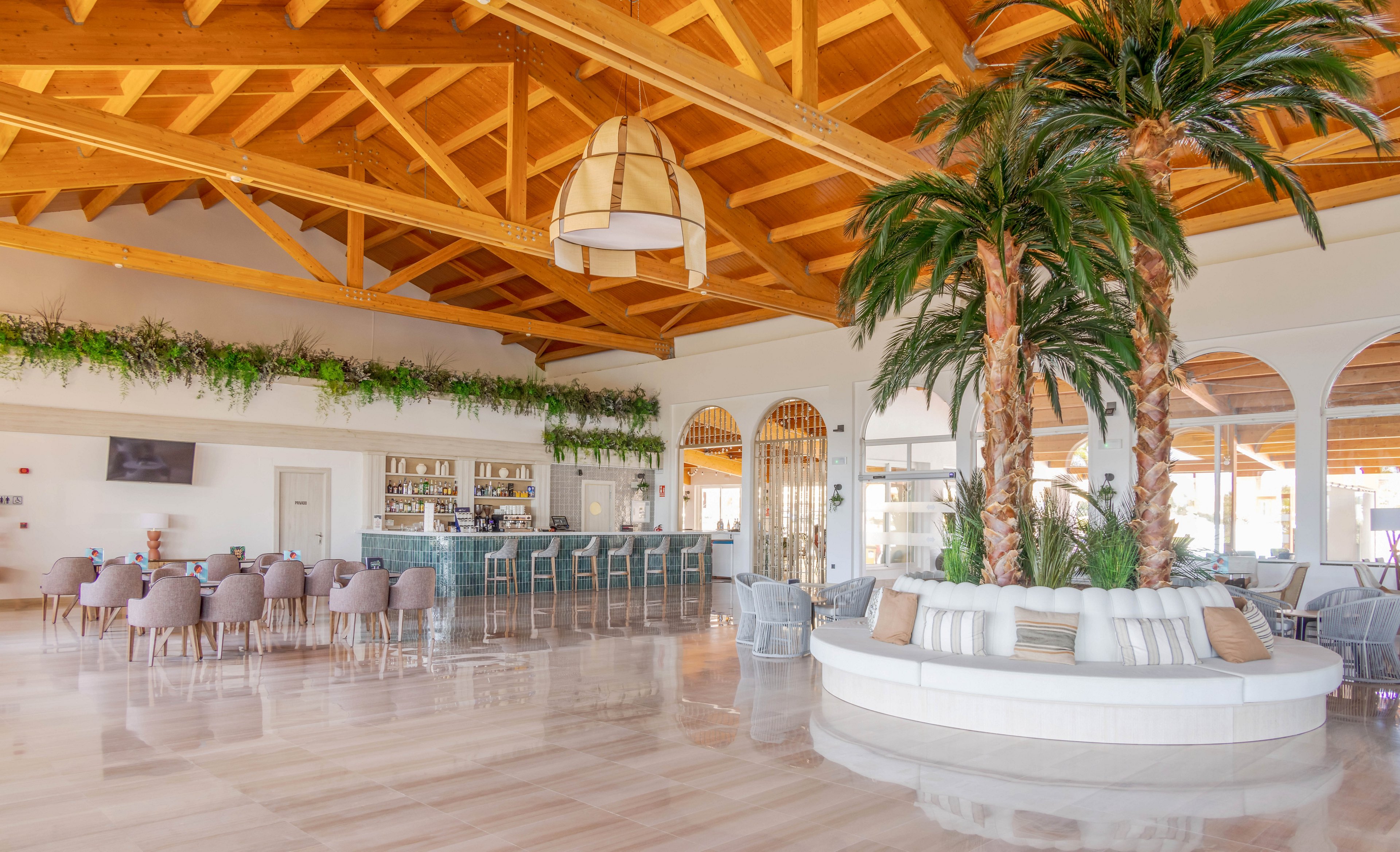 A bright, airy lounge bar features a central seating area with tall indoor palm trees and plush cushions. The bar area is lined with blue chairs and backed by shelves of bottles, while greenery decorates the upper walls. The space is illuminated by natural light and a large woven pendant lamp, with a high wooden ceiling adding warmth. The overall palette is neutral with touches of green and blue, creating a relaxed, contemporary atmosphere.