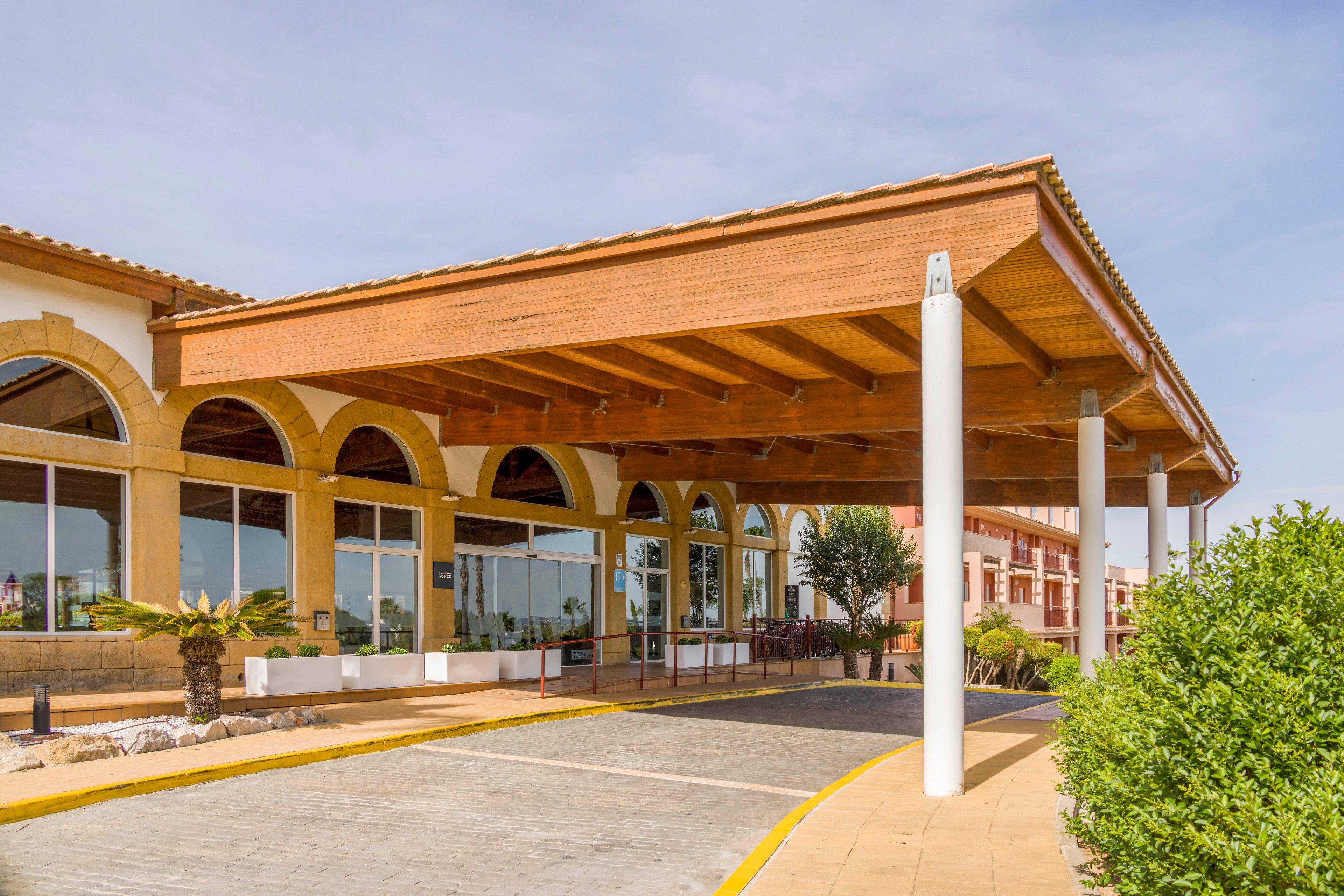 A contemporary hotel entrance features a large wooden canopy supported by white columns. The building showcases arched windows and a clean, paved driveway leading to the entrance. The setting is outdoors on a sunny day, with greenery and landscaping visible around the structure. The overall mood is welcoming and professional, with warm tones and clear skies.