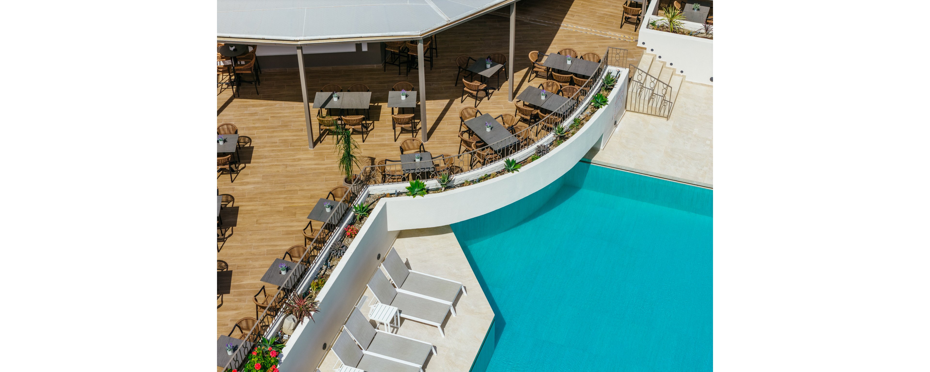 A bright hotel pool area is viewed from above, featuring a curved white deck beside clear turquoise water. Several white sun loungers line the pool edge, accented by planters with green foliage and red flowers. Beyond the pool, a wooden terrace holds multiple tables and chairs under a partial canopy, suggesting an outdoor dining or lounge space. The scene feels clean, modern, and inviting, ideal for leisure and relaxation in warm weather.