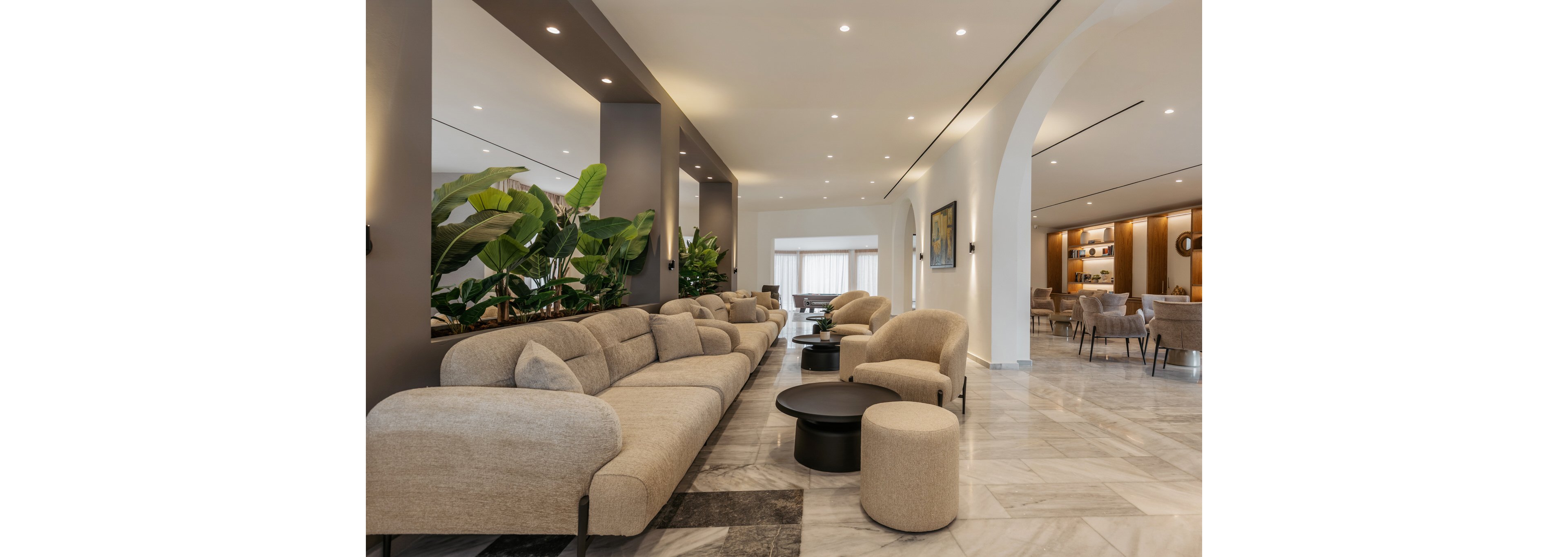 A spacious contemporary lobby features long beige sofas and rounded armchairs arranged around low circular coffee tables. Large green indoor plants add a natural accent beside the seating area, while recessed ceiling lights create an even, welcoming illumination. Marble flooring and tall columns emphasize the clean, modern architecture, leading toward an arched opening into a dining or lounge space in the background. The overall atmosphere is calm, elegant, and designed for comfortable socializing or relaxation.
