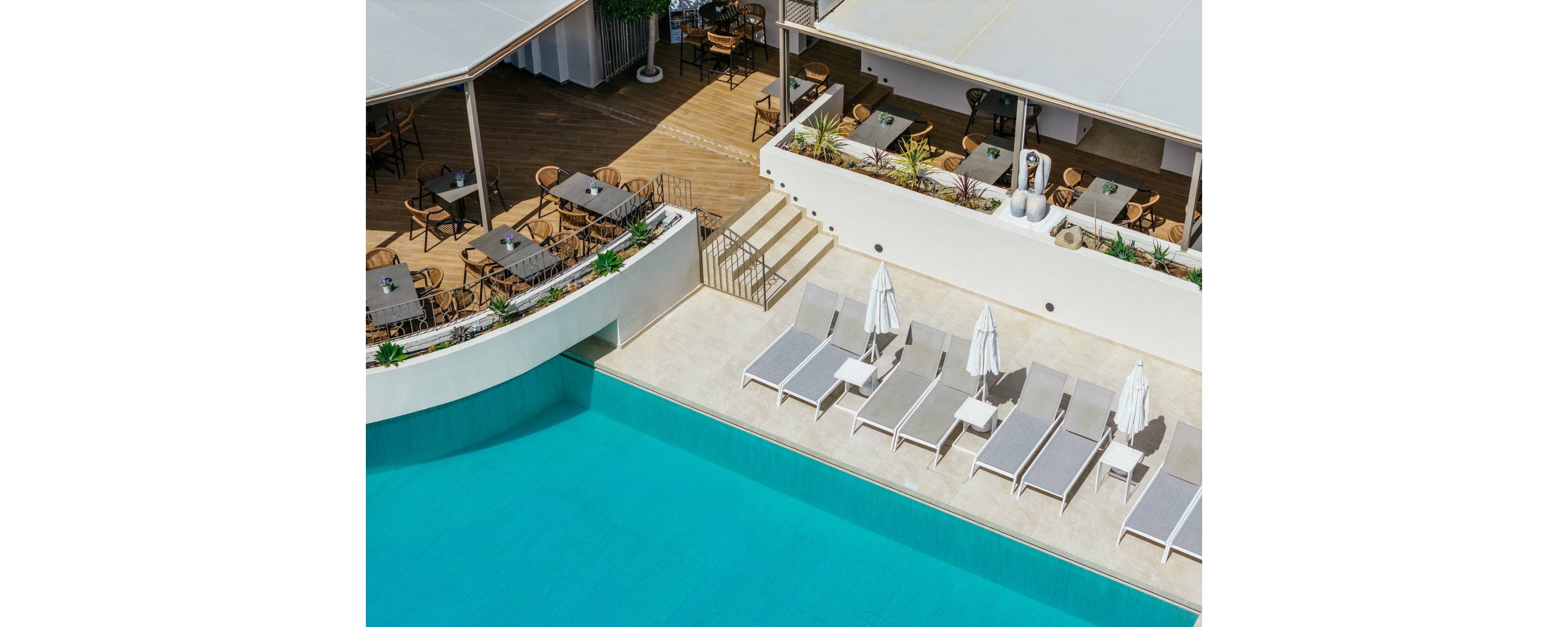 A bright turquoise swimming pool sits beside a row of white sun loungers with umbrellas. Above the pool, an open-air terrace features wooden tables and chairs arranged for outdoor dining. The setting appears clean and modern, with light-colored walls and minimal decor. Strong sunlight and shadows create a relaxed vacation atmosphere.