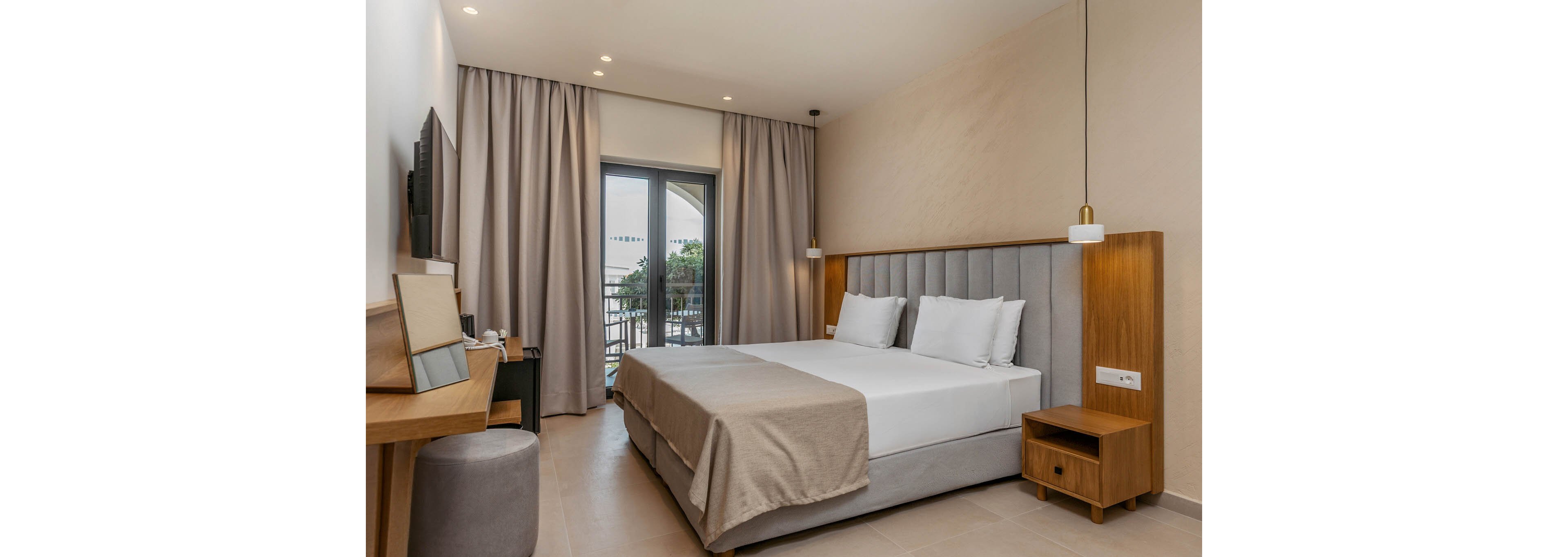 Contemporary hotel room featuring a large double bed with neutral bedding and upholstered headboard. The space includes a wooden nightstand, desk, and wall-mounted television, creating a functional guest environment. Floor-to-ceiling curtains frame a glass door leading to a balcony with outdoor greenery. Soft, warm lighting and beige tones contribute to a calm, minimalist atmosphere.
