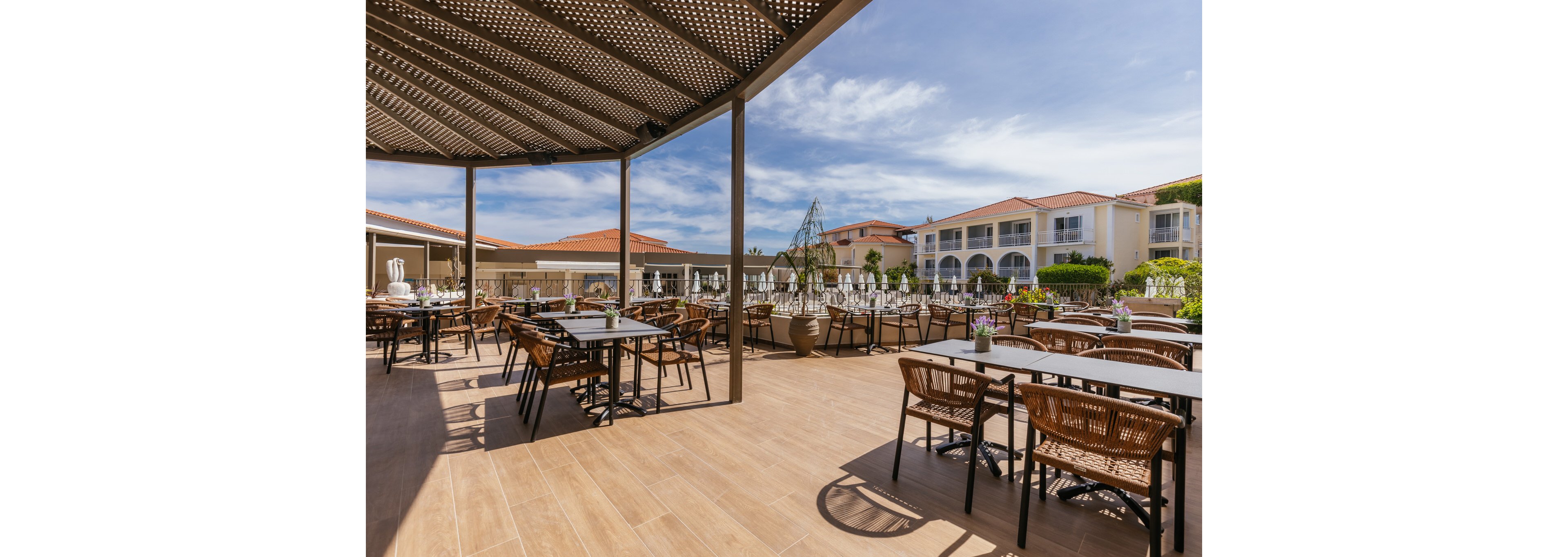 Spacious outdoor dining terrace at a resort with neatly arranged tables and wicker chairs. The setting features a partial pergola roof casting patterned shadows on the light wooden floor. In the background, white multi-story resort buildings with red-tiled roofs frame the scene under a bright blue sky with scattered clouds. Decorative plants and table settings add a relaxed, vacation atmosphere to the open-air restaurant space.