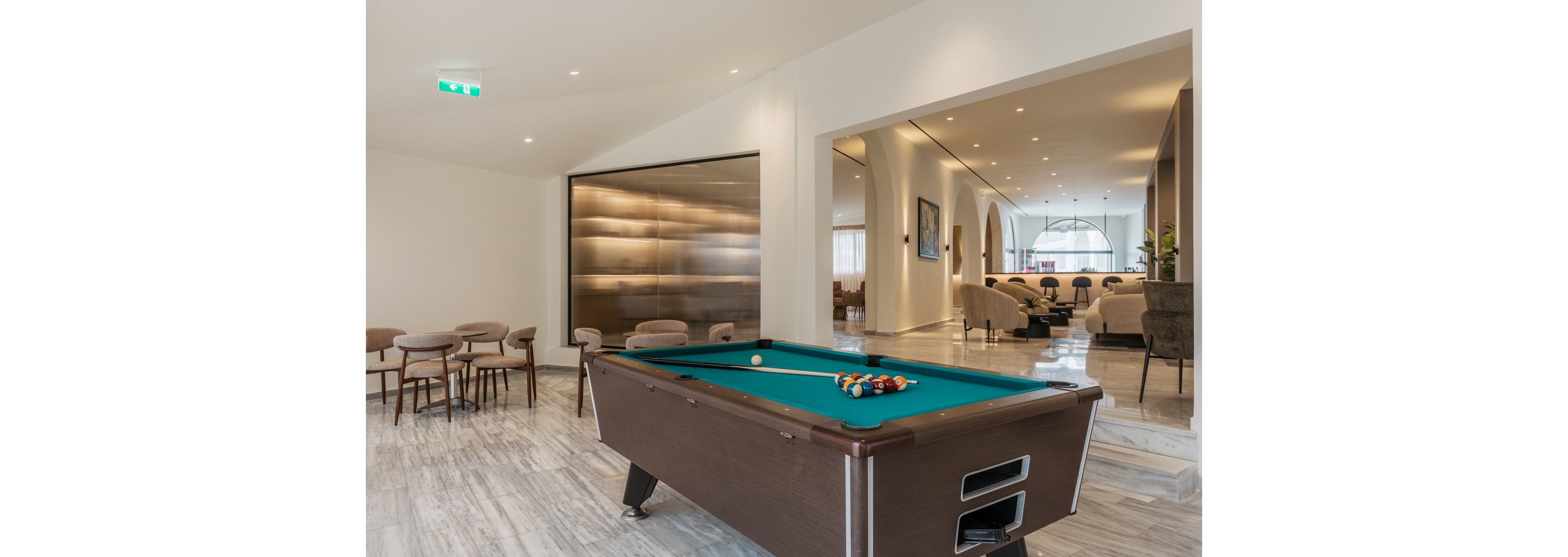A contemporary lounge features a dark wooden billiard table as the central focus. The space has light marble flooring, neutral walls, and minimalist furniture including small tables and chairs. In the background, an open seating area with armchairs and soft lighting creates a relaxed social atmosphere. Overall, the interior feels spacious, elegant, and designed for leisure and conversation.