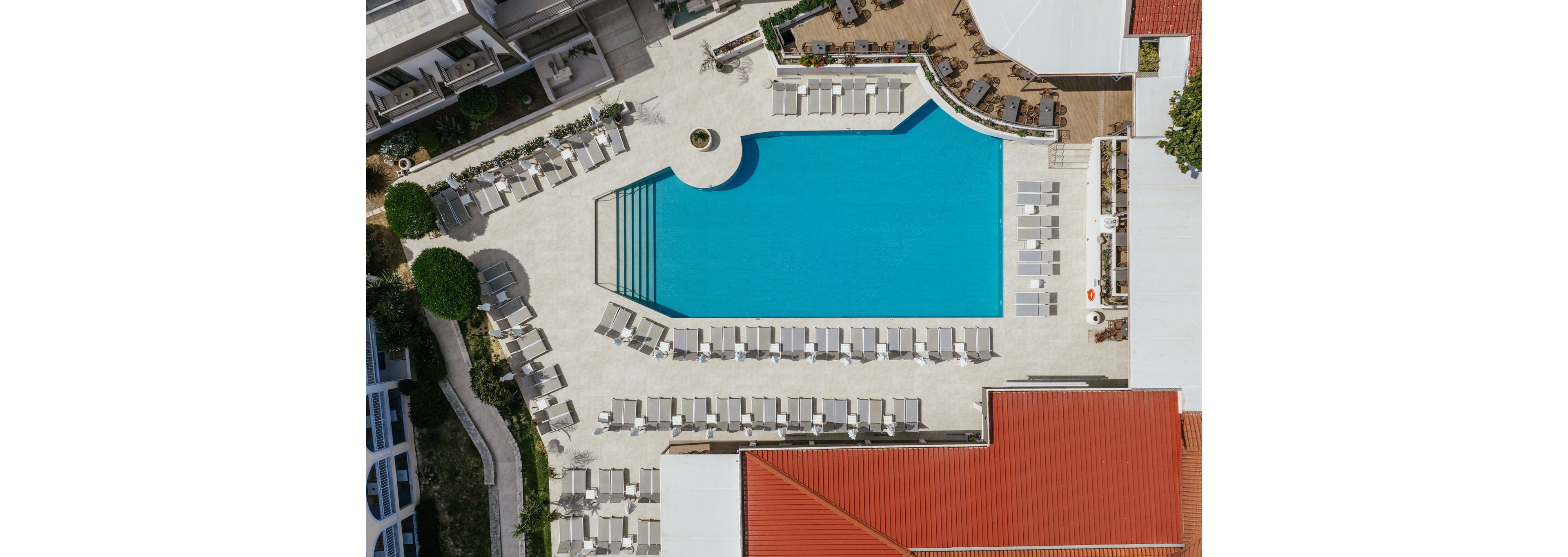 A bright blue rectangular swimming pool is seen from above in a contemporary hotel or resort courtyard. Rows of white sun loungers and umbrellas surround the pool on a light-colored terrace, creating a clean, organized layout. Stair steps lead into the water at one end, and landscaped greenery borders parts of the area. Nearby buildings with red and white roofs frame the pool, emphasizing the geometric design of the space.