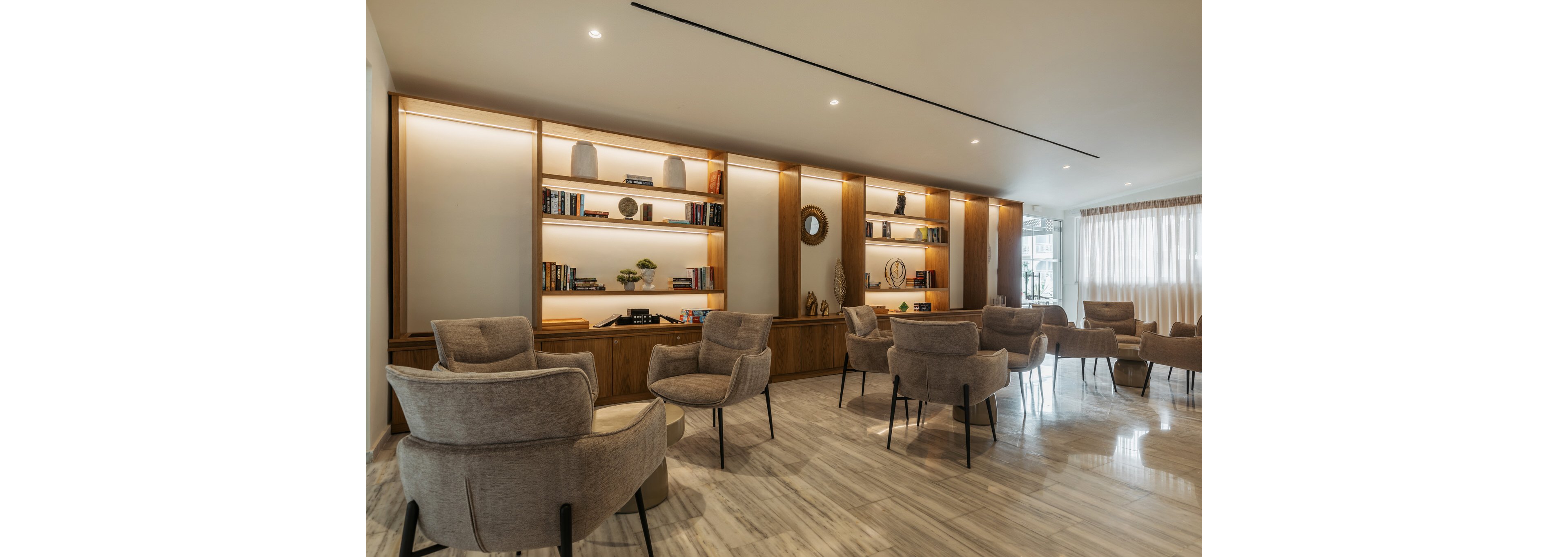 A contemporary lounge area features multiple cushioned armchairs arranged for conversation. Tall wooden bookshelves line the back wall, displaying books, vases, and decorative objects. Recessed ceiling lights and integrated shelf lighting create a warm, inviting atmosphere. Large windows with sheer curtains allow natural light to brighten the polished floor and neutral-toned furnishings.