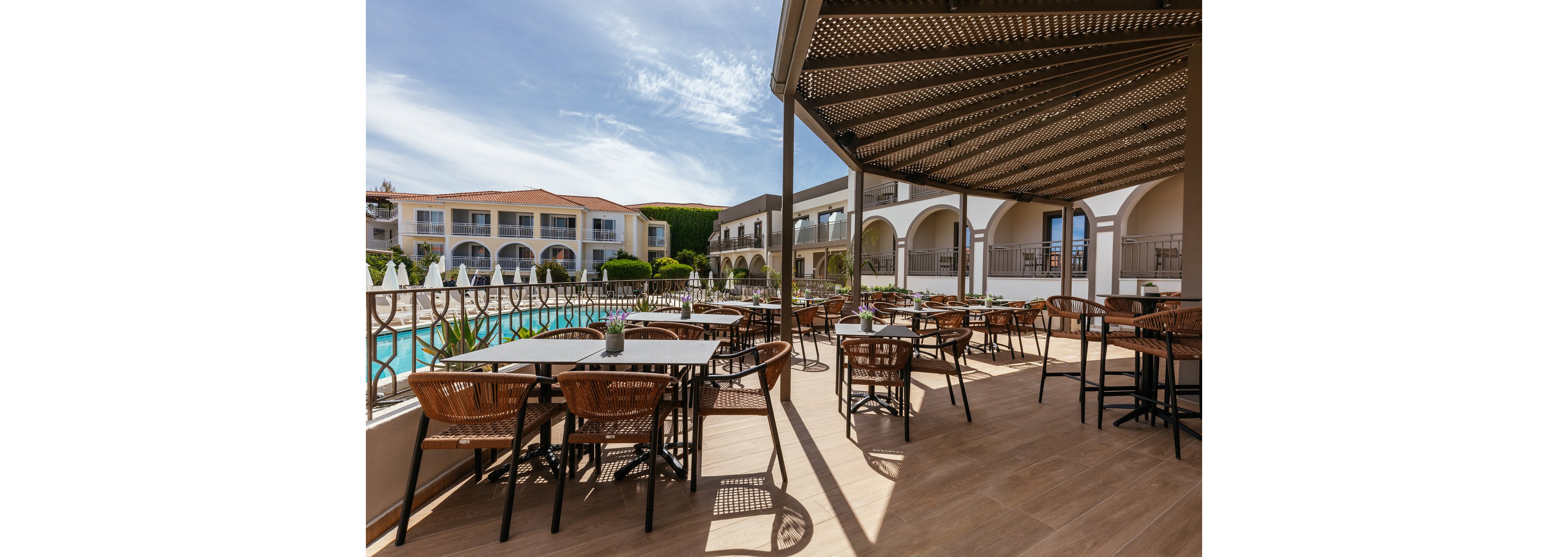 An open-air dining terrace overlooks a large swimming pool at a Mediterranean-style resort. Wicker chairs and dark tables are neatly arranged under a partial pergola providing shade. In the background, white buildings with arches and balconies frame the courtyard setting. Potted plants and clear blue sky add a relaxed vacation atmosphere.