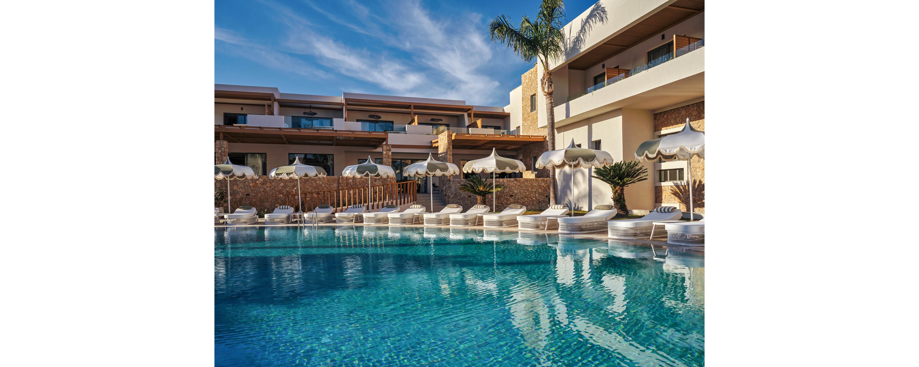 A luxurious resort pool glistens under clear blue skies in front of a modern hotel building. White sun loungers and parasols line the poolside, creating an inviting relaxation area. Contemporary architecture with stone accents and balconies frames the scene. Palm trees and manicured plants add a tropical touch to the tranquil outdoor setting.