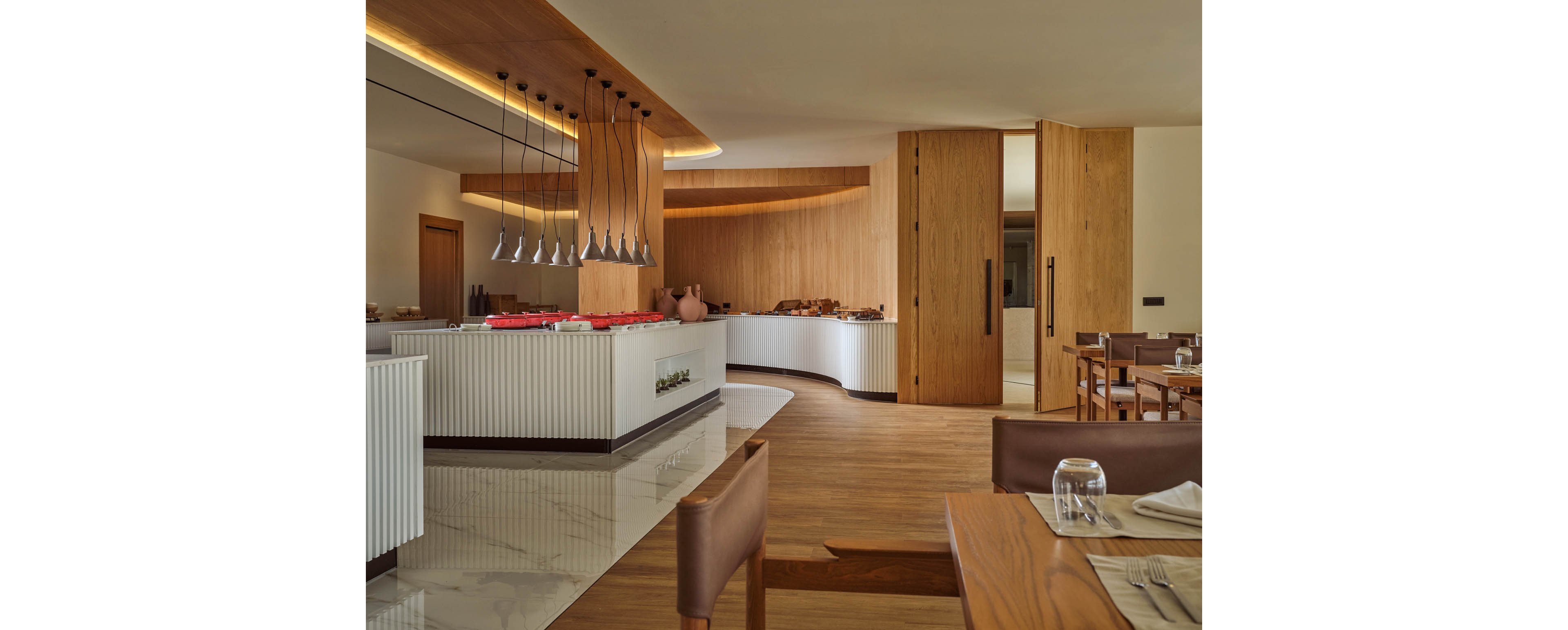 A contemporary buffet area features sleek white counters and warm wooden finishes. Pendant lights hang above the serving station, highlighting neatly arranged dishes and tableware. The space opens into a dining area with set tables, leather chairs, and folded napkins. Soft, indirect lighting and clean lines create a calm, upscale restaurant atmosphere.