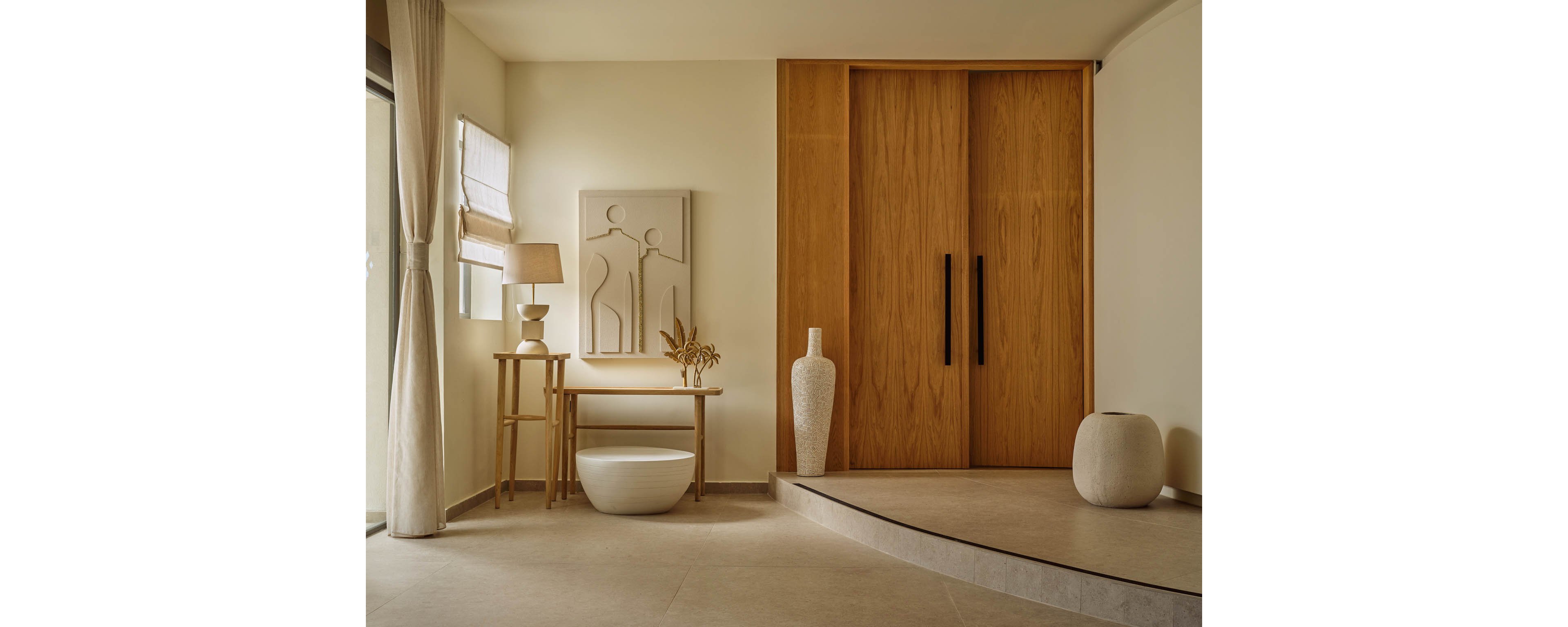 A minimalist living space features large wooden sliding doors as the focal point. The room is decorated in warm neutral tones with a small side table, lamp, and sculptural wall art. Ceramic vases and a low round bowl add organic shapes to the clean interior. Soft natural light enters through a curtained window, enhancing the calm and serene atmosphere.