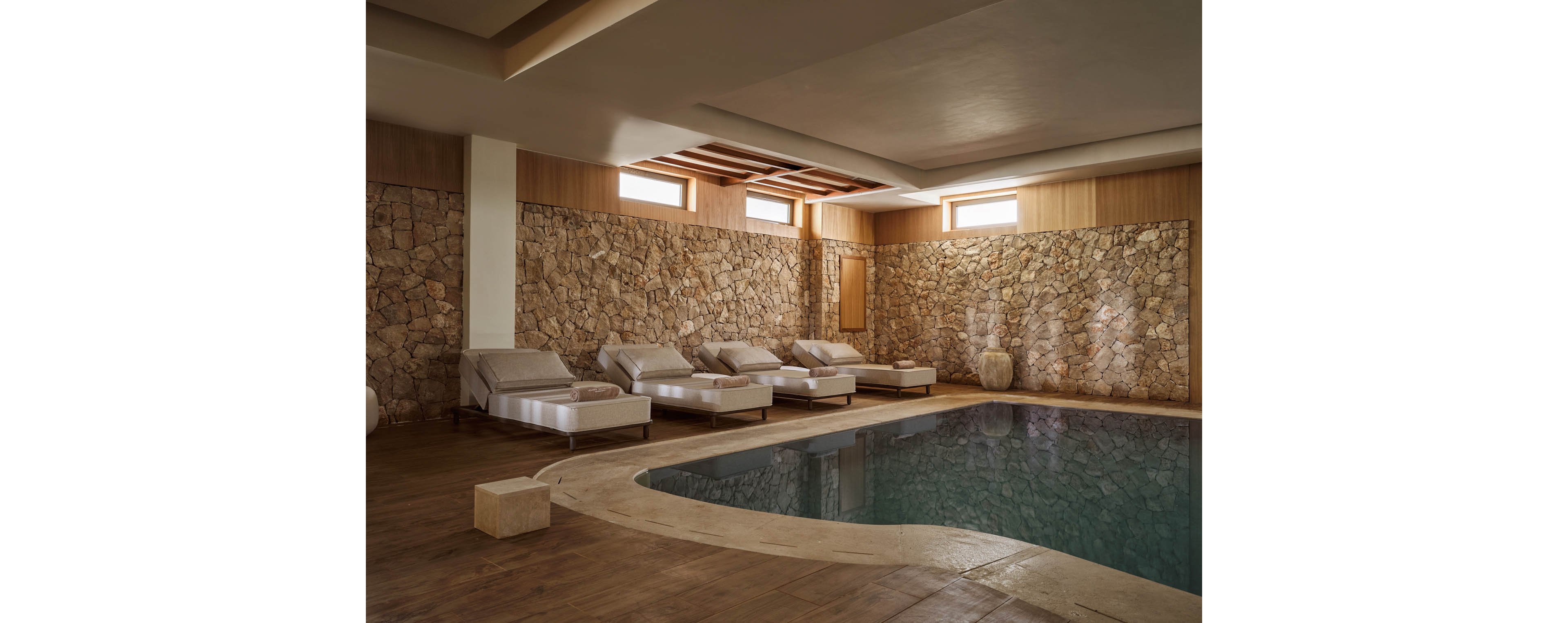 An indoor spa area features a curved pool surrounded by cushioned lounge chairs. The space is enclosed by textured stone walls and warm wooden paneling. Soft natural light enters through high rectangular windows, creating gentle reflections on the water. The overall atmosphere is calm and inviting, ideal for relaxation and wellness.