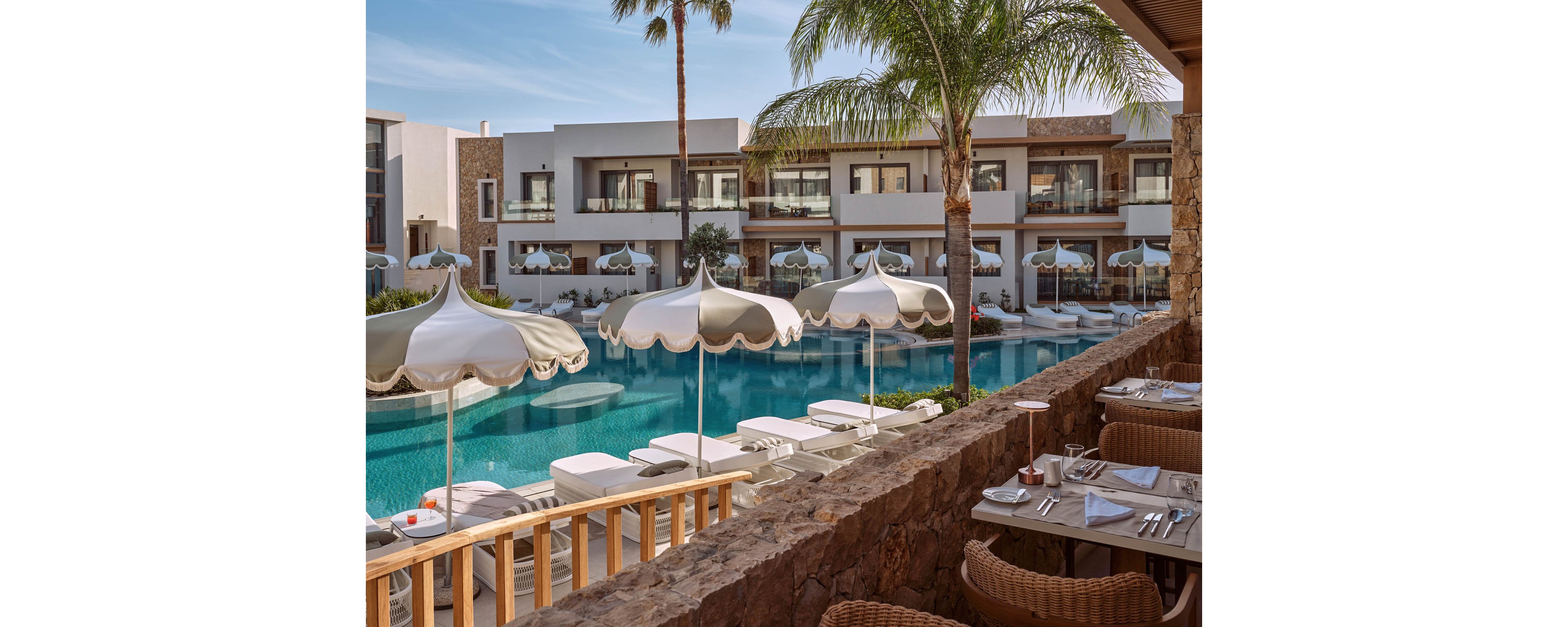 A stylish resort features a large swimming pool surrounded by white sunbeds and decorative parasols. In the foreground, an outdoor dining area is set with white tableware and wicker chairs along a stone terrace. Modern two-story suites with balconies overlook the pool, framed by palm trees and clear skies. The scene conveys a relaxed, upscale vacation atmosphere in a warm, sunny climate.