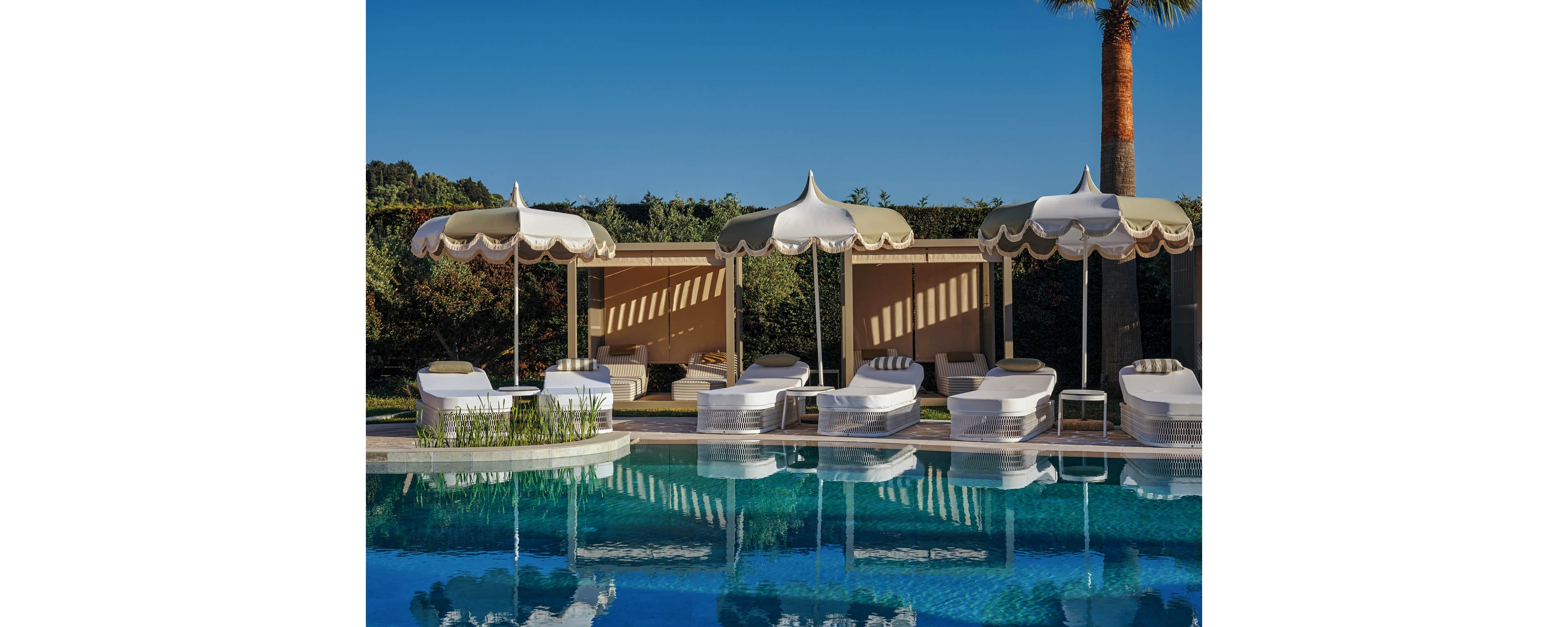 A row of white cushioned loungers lines the edge of a clear blue swimming pool. Elegant scalloped sun umbrellas provide shade in front of private cabana-style seating areas. Lush greenery and a tall palm tree frame the serene outdoor setting under a bright blue sky. The scene evokes a luxurious, tranquil resort atmosphere ideal for relaxation and leisure.