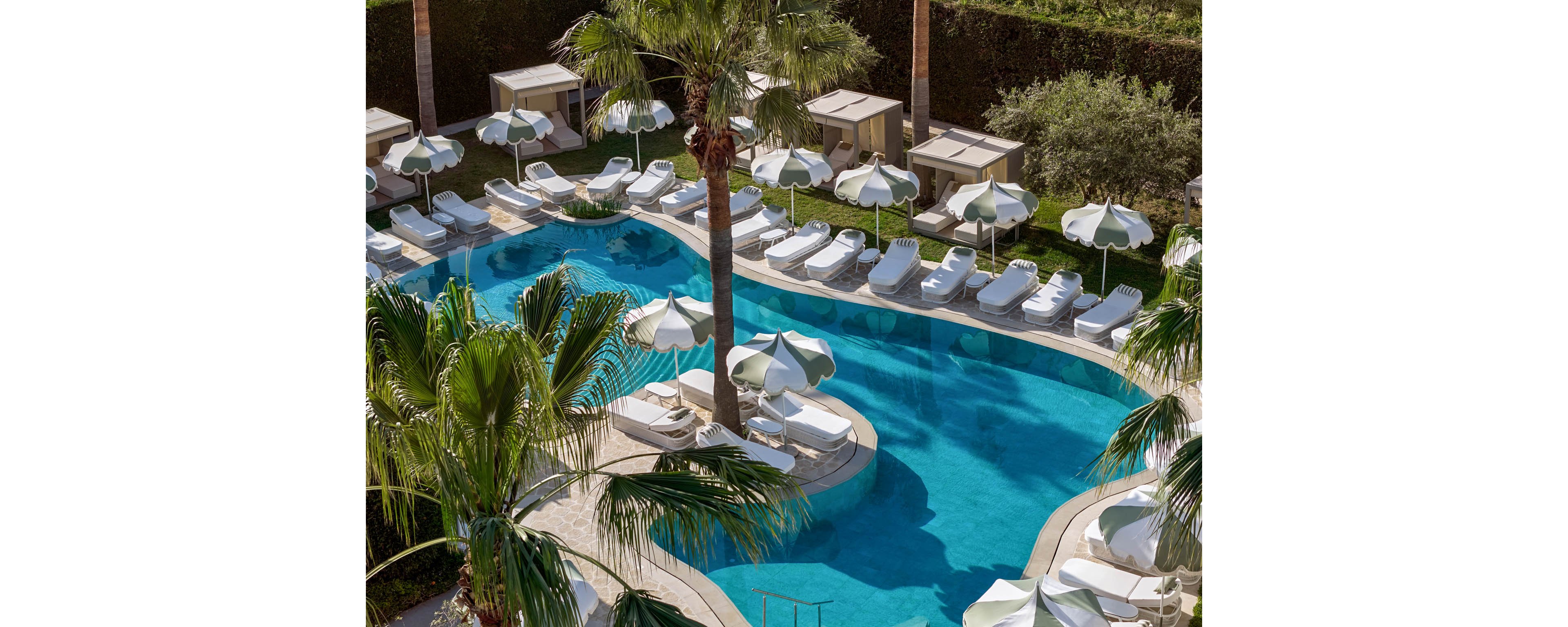 Curved turquoise swimming pools are surrounded by numerous white sun loungers and matching umbrellas. The scene appears to be a luxury resort or hotel outdoor area, viewed from above. Palm trees and lush greenery frame the pool deck, adding a tropical atmosphere. Private cabanas line the back edge, emphasizing relaxation and vacation leisure.