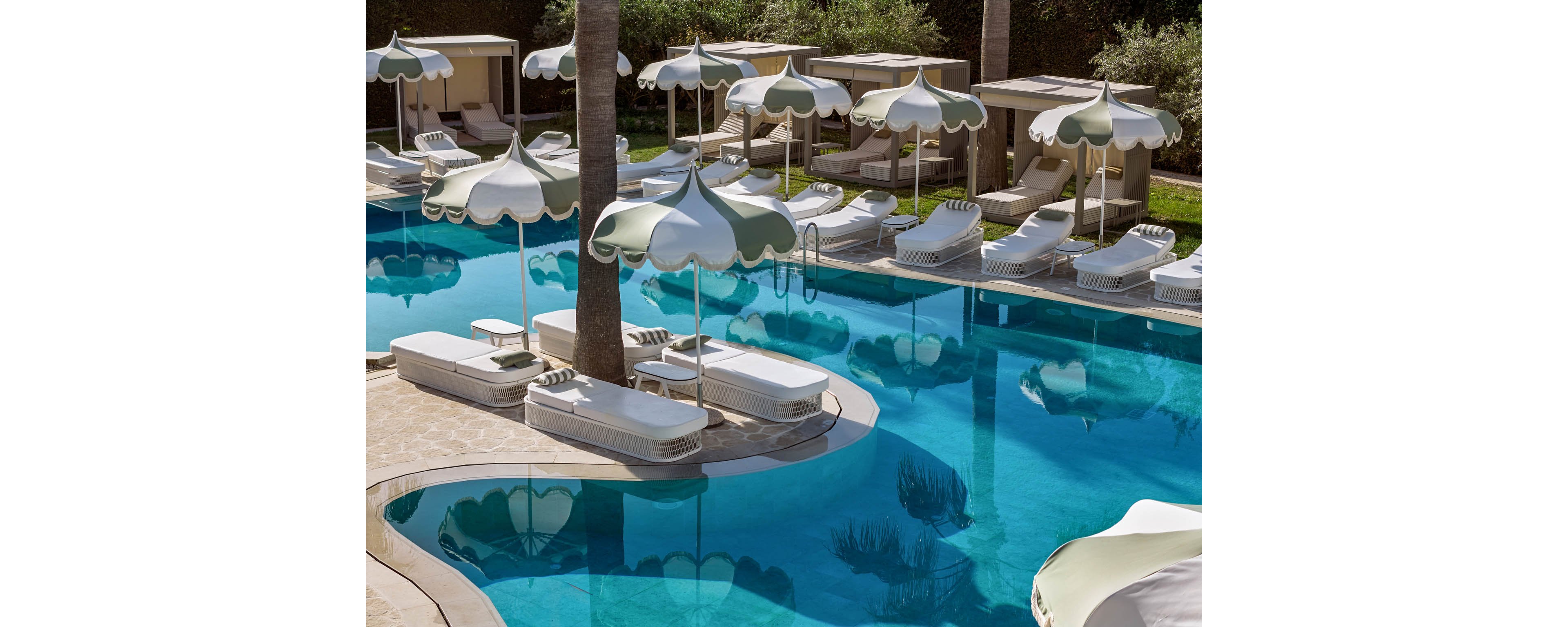 A tranquil resort swimming pool is lined with white cushioned sun loungers and cabanas. Curved pool edges and clear blue water create an inviting, upscale atmosphere. White and green scalloped umbrellas provide shade along the poolside deck. The scene is empty of people, emphasizing relaxation and exclusivity in a sunny outdoor setting.