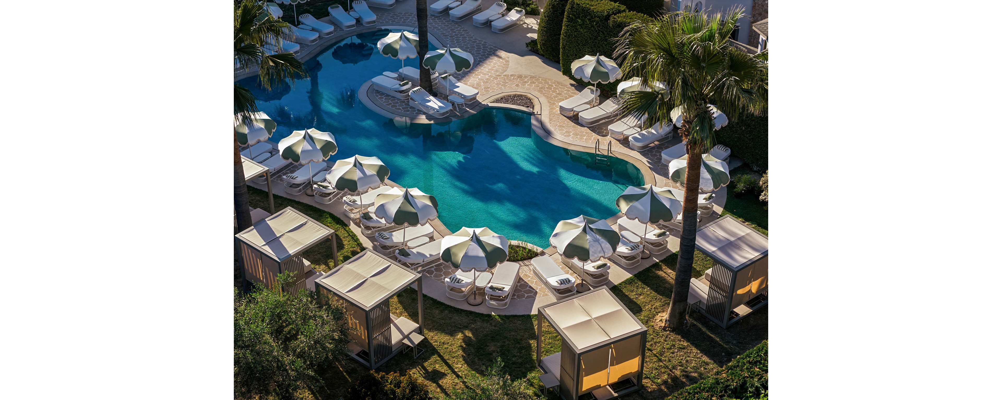 An inviting resort swimming pool is surrounded by white lounge chairs and striped umbrellas. Private cabanas with fabric canopies line the grassy edge, offering shaded relaxation areas. Tall palm trees and manicured hedges frame the curving pool, creating a secluded oasis. The scene is sunlit and tranquil, suggesting a warm vacation atmosphere.