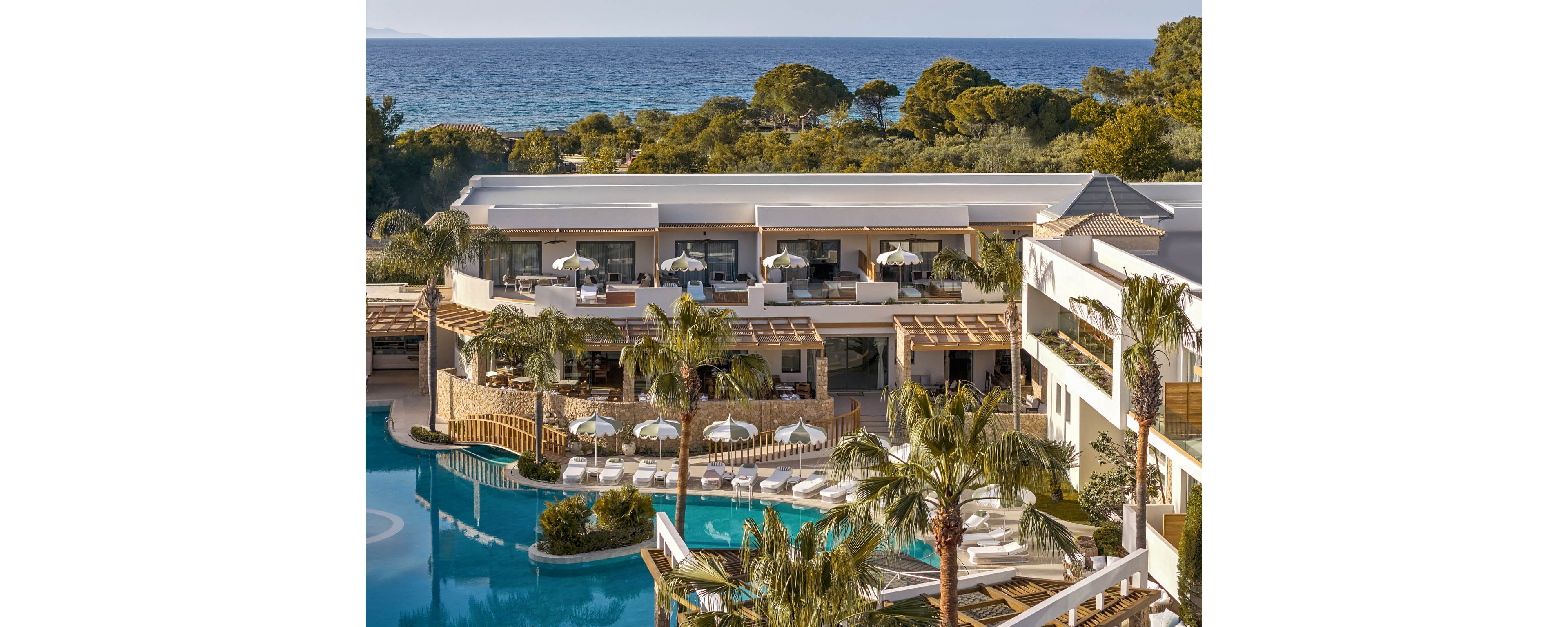 A modern luxury resort overlooks a large curving swimming pool surrounded by palm trees and sun loungers. White umbrellas and shaded terraces create inviting outdoor relaxation areas. The multi-story buildings feature balconies and wooden pergolas facing the water. In the background, the calm sea and distant shoreline complete the tranquil coastal setting.