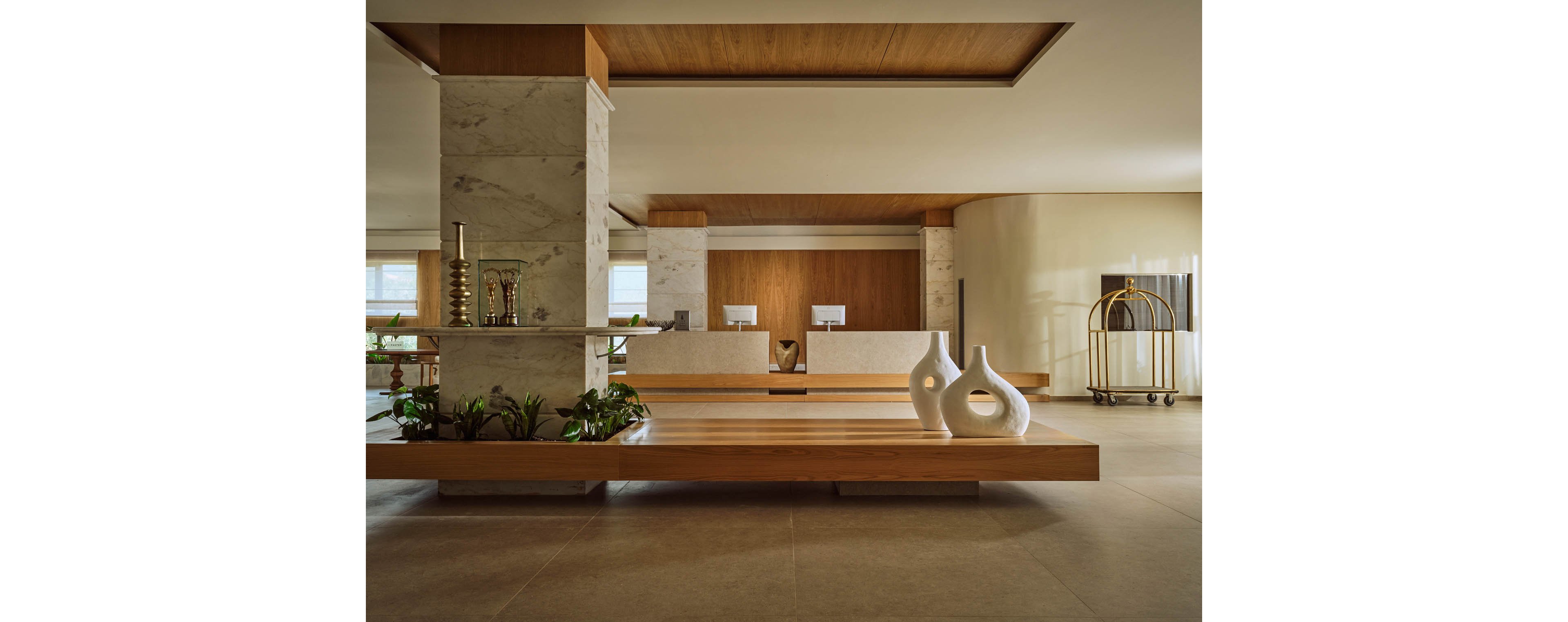 A contemporary hotel lobby features a long wooden bench and minimalist reception desk. The space is accented with marble columns, warm wood paneling, and neutral-toned flooring. Sculptural white vases and green plants add decorative interest to the clean, open layout. A brass luggage cart stands to the side, emphasizing the hospitality setting.