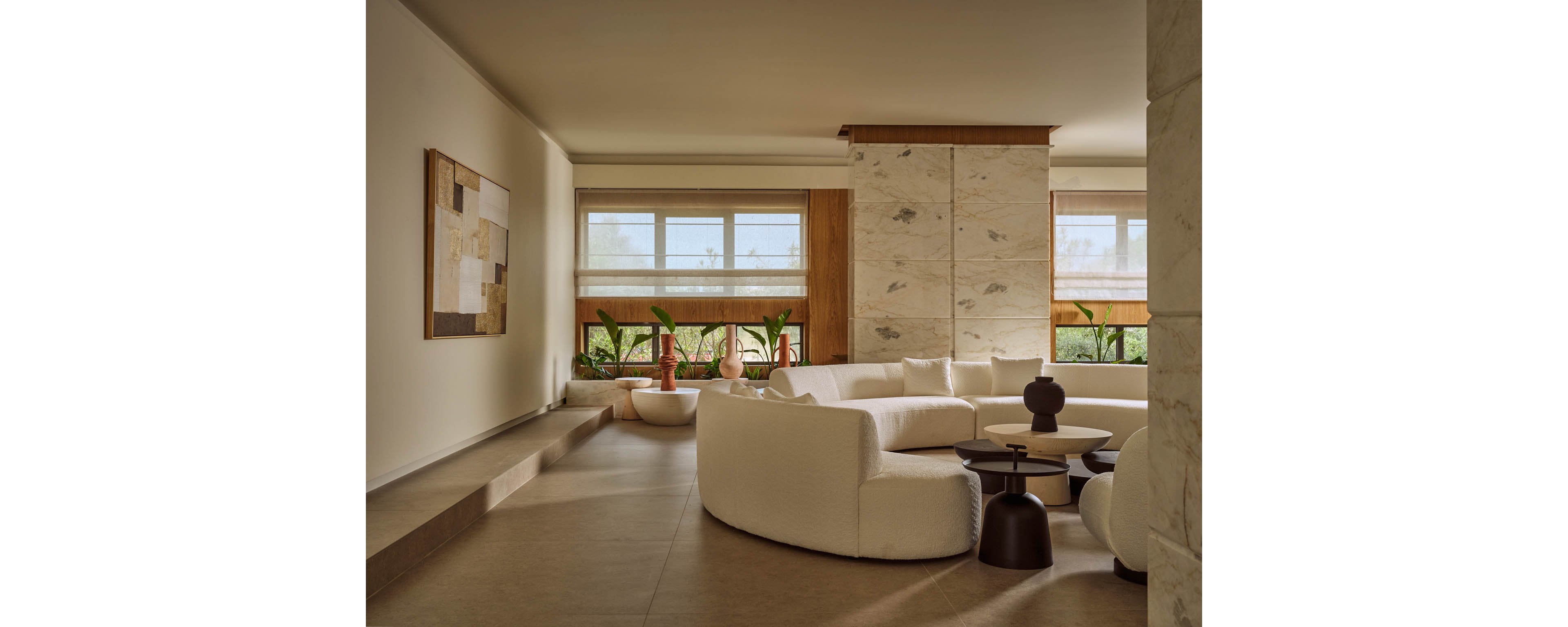 A contemporary living room features a large curved white sofa arranged around low circular coffee tables. The space is defined by neutral tones, smooth stone flooring, and a tall marble-clad column. Large windows with light blinds allow natural daylight to illuminate indoor plants along the wall. A geometric abstract artwork hangs on the left, enhancing the modern minimalist aesthetic.