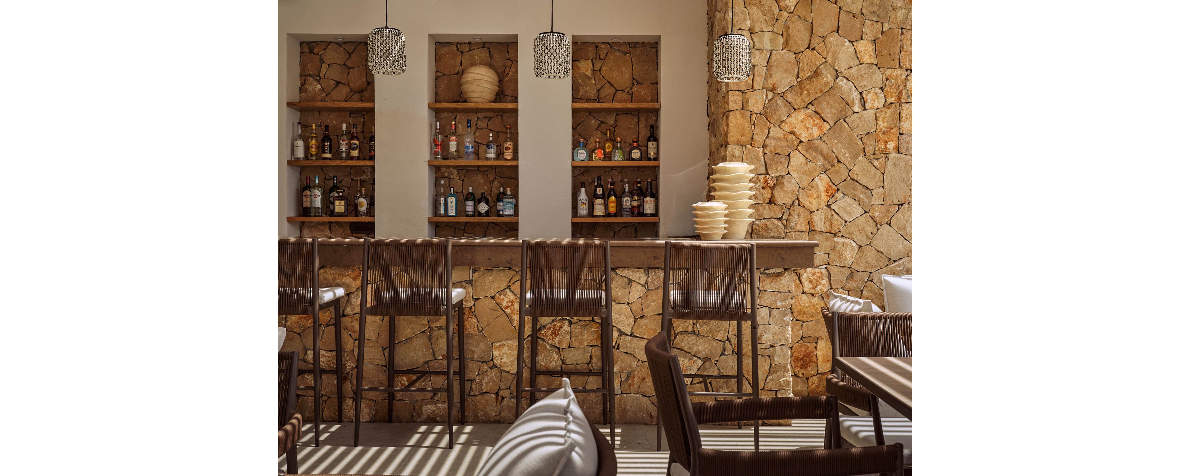 A stylish bar with a stone wall backdrop features wooden shelves lined with assorted liquor bottles. Tall bar stools with woven backs are neatly arranged along a smooth wooden counter. Pendant lights hang from the ceiling, casting soft shadows across the seating area. Stacks of light-colored bowls sit on the counter, adding a subtle decorative touch to the warm, sunlit interior.