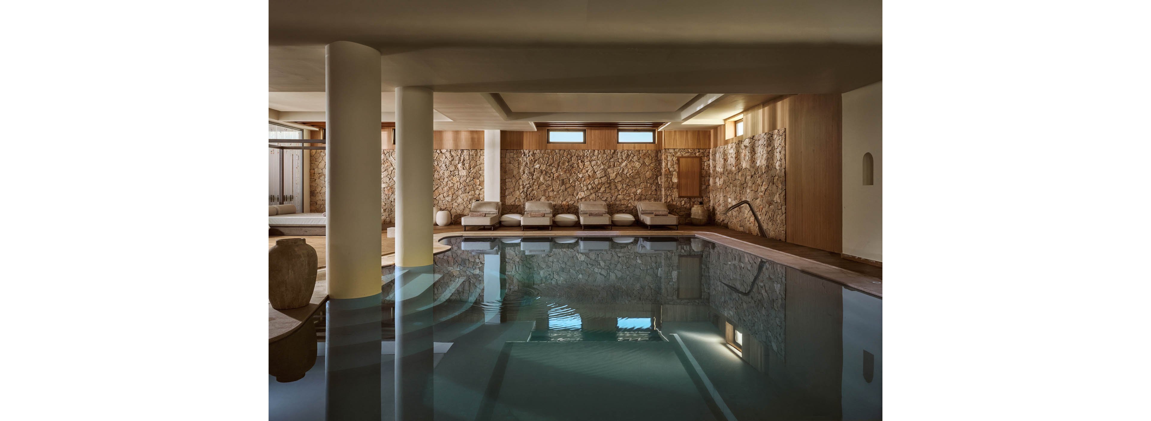 A tranquil indoor swimming pool is surrounded by cushioned lounge chairs against a textured stone wall. Soft natural light filters through small high windows, creating gentle reflections on the calm water. Warm wood paneling and neutral tones contribute to a minimalist, luxurious spa atmosphere. A metal handrail leads into the pool, inviting relaxation and quiet retreat.