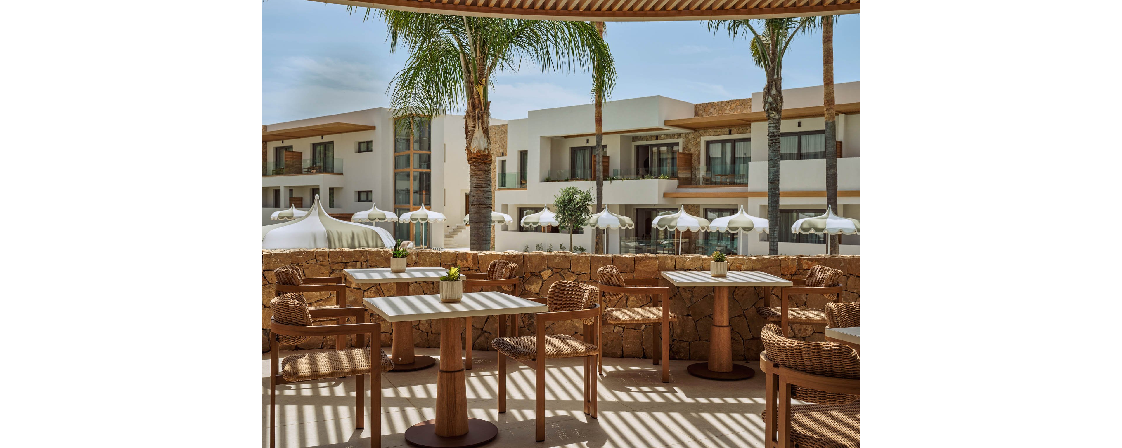 An outdoor terrace cafe features wooden tables and woven chairs arranged neatly under a shaded pergola. Palm trees rise between the seating area and a row of modern resort buildings with balconies. White umbrellas and cabana-style tents add a relaxed, vacation atmosphere. Warm neutral tones and natural materials create a calm, Mediterranean-inspired setting.