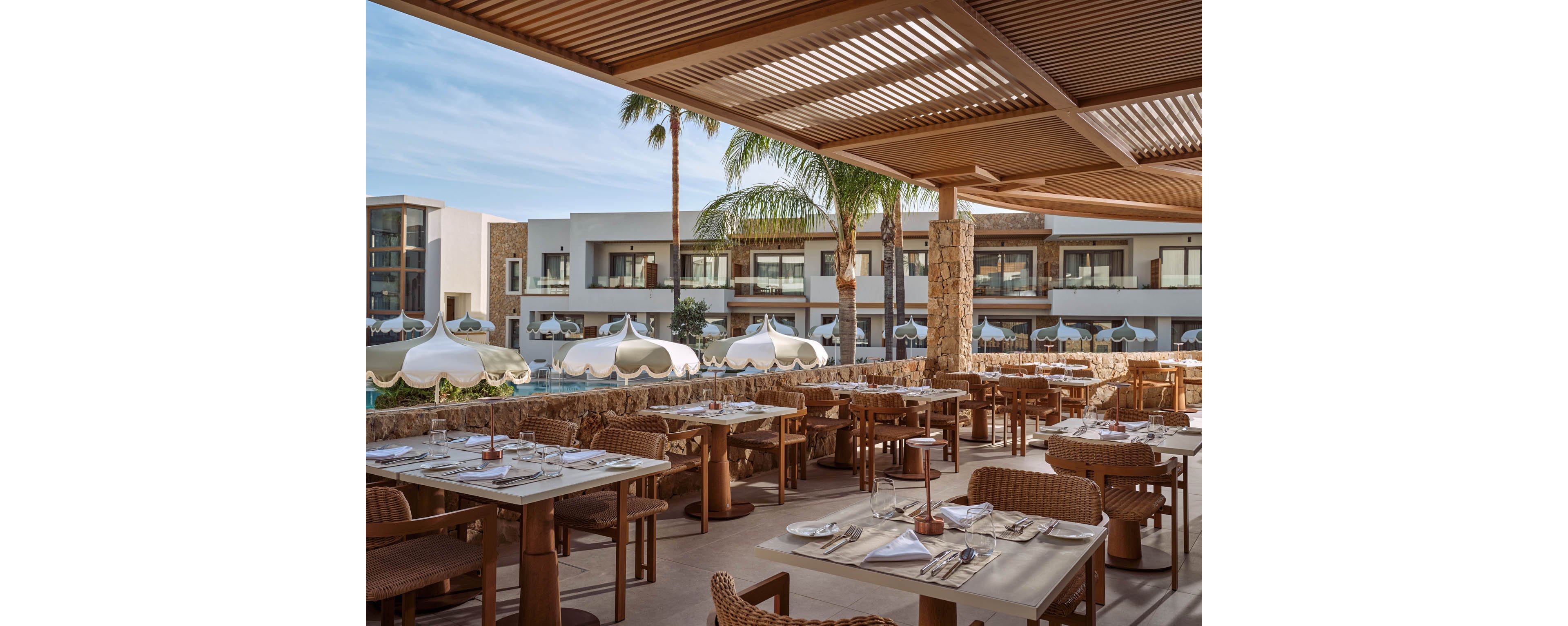 An elegant open-air restaurant terrace overlooks a modern resort courtyard. Neatly set tables with woven chairs face a row of white poolside cabanas and sun loungers. Palm trees and contemporary low-rise buildings frame the relaxed vacation setting. Warm natural tones and soft daylight create a calm, upscale holiday atmosphere.