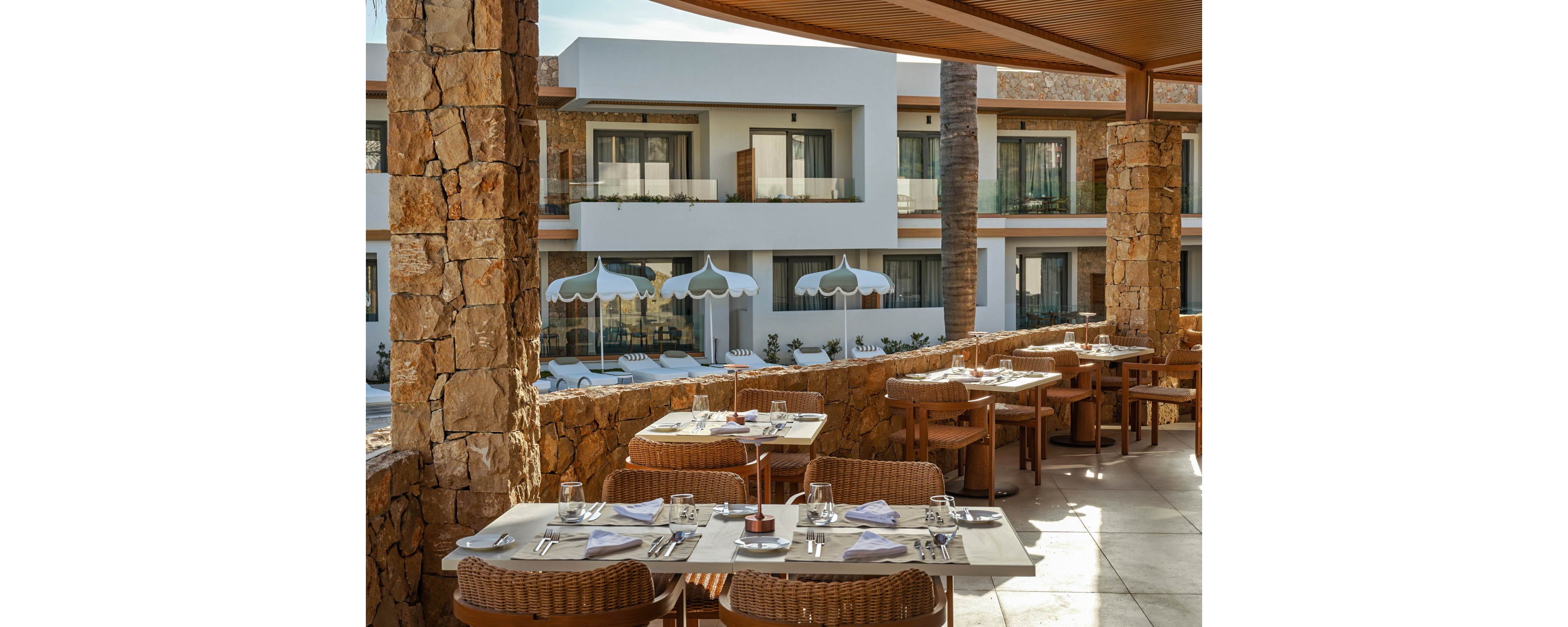 An elegant outdoor restaurant terrace is set with neatly arranged tables, glassware, and white napkins. The dining area features wicker chairs and stone columns under a wooden roof. In the background, a modern hotel building with balconies faces a pool area lined with white sun loungers and scalloped umbrellas. Warm natural light enhances the relaxed, upscale vacation atmosphere.